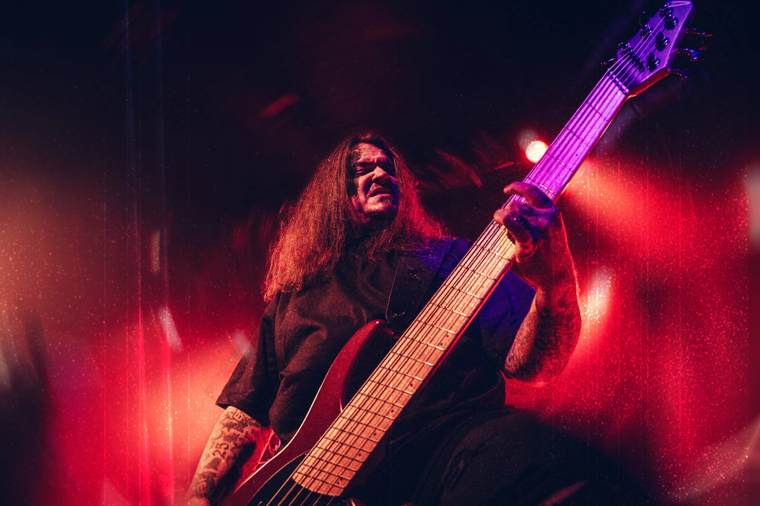 Ein Bassist auf der Bühne umhüllt mit roten verwirbeltem Licht. Er guckt besonders grimmig. Das Bild wirkt aufgeladen.