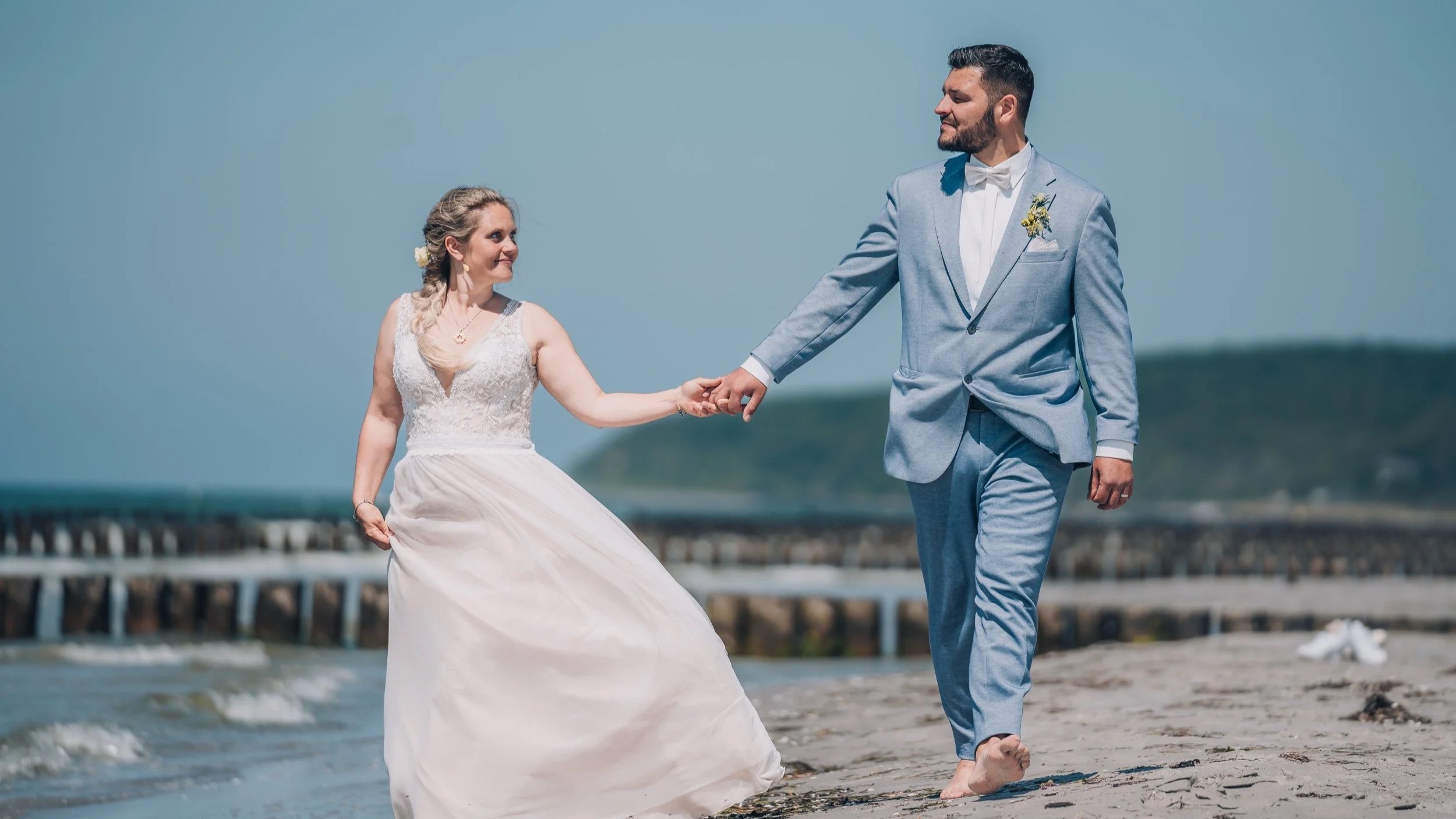 Ein Brautpaar in formeller Kleidung beim Spaziergang am Strand, die Frau in Brautkleid, der Mann in Anzug, bei schönem Wetter im Freien.