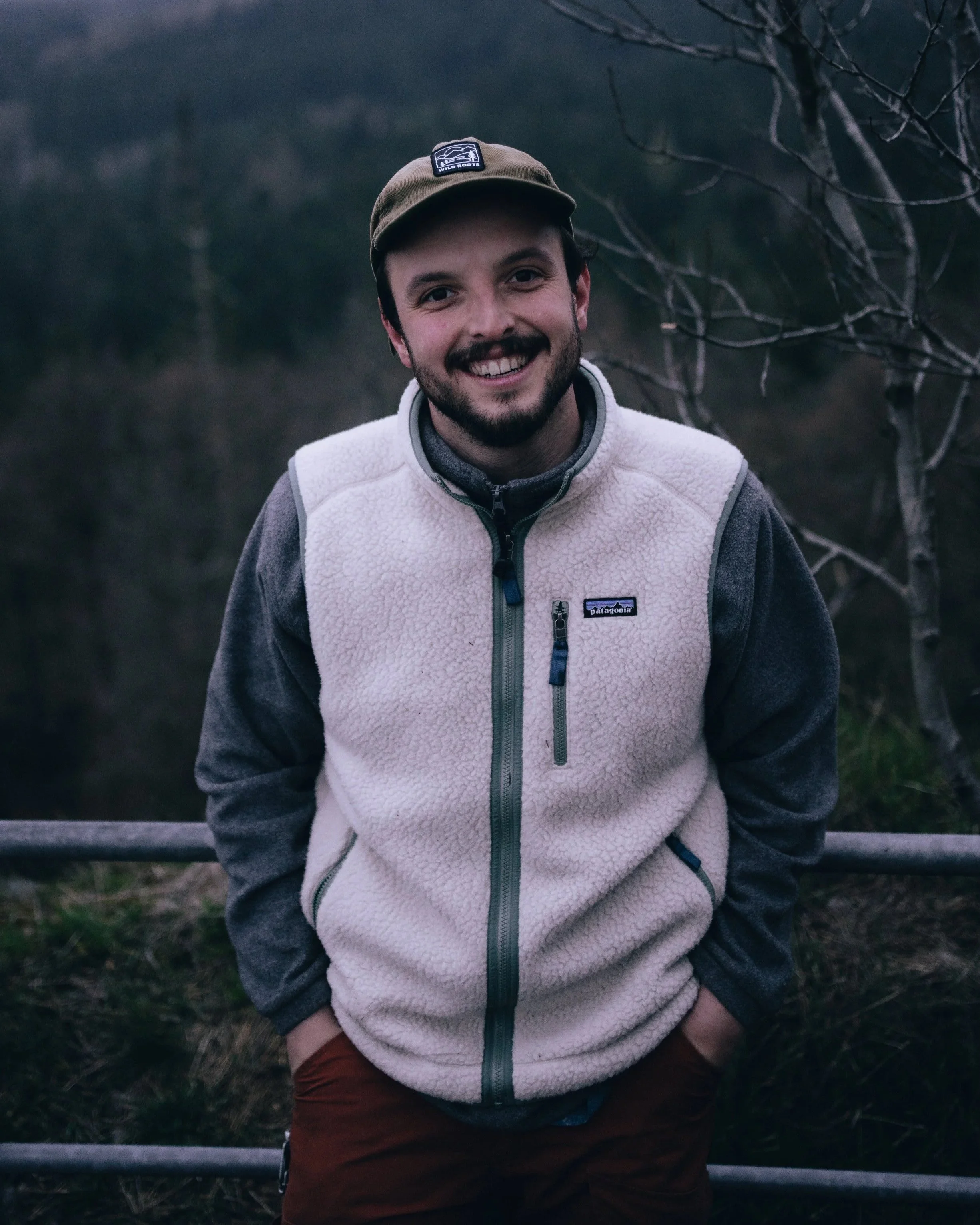 Ein lachender Mann trägt eine beige fleece Weste und eine braune Kappe, im Freien mit Bäumen im Hintergrund.