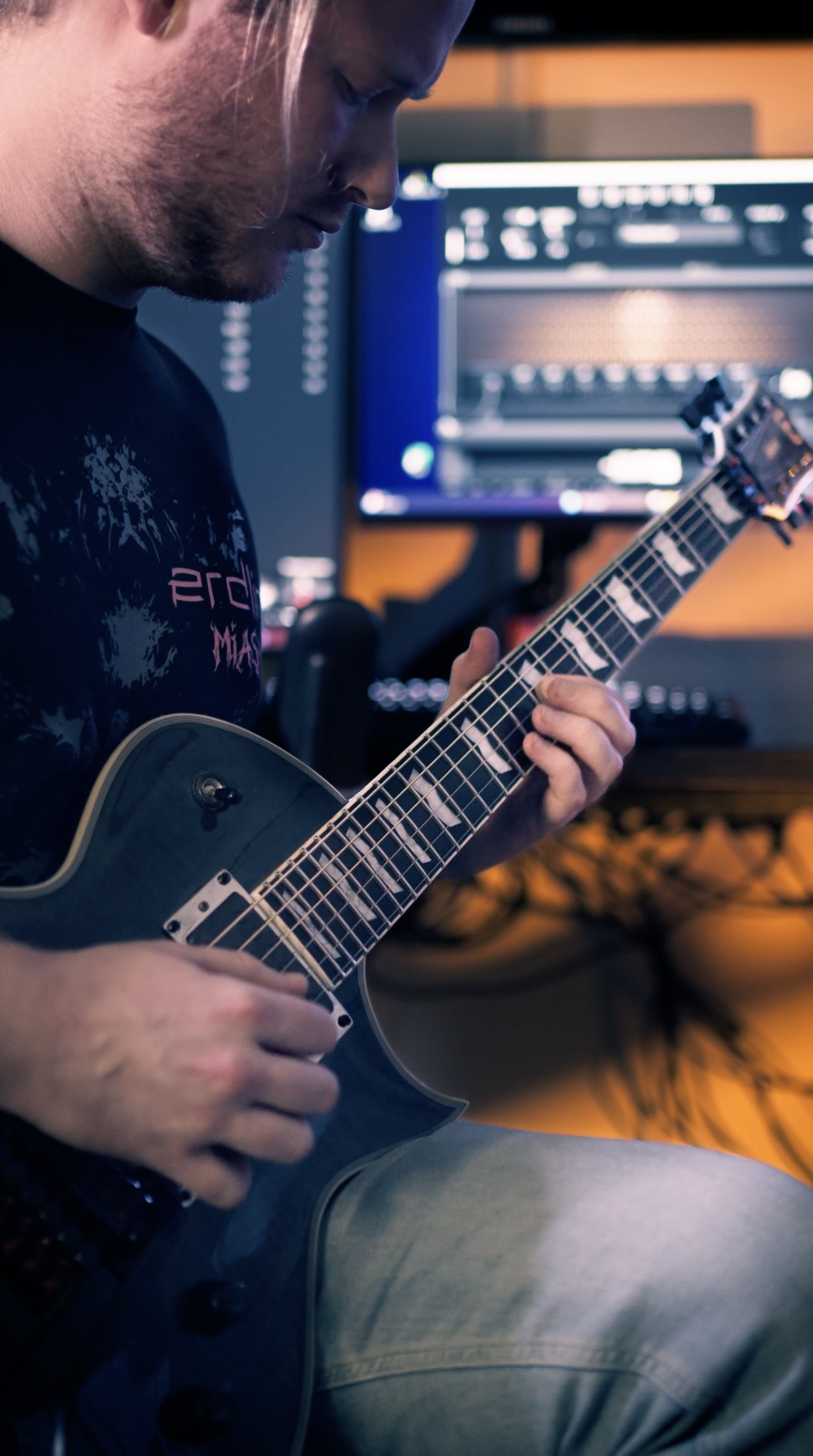 Ein Mann spielt Gitarre in einem Tonstudio, im Hintergrund sind Computerbildschirme mit Musiksoftware zu sehen.