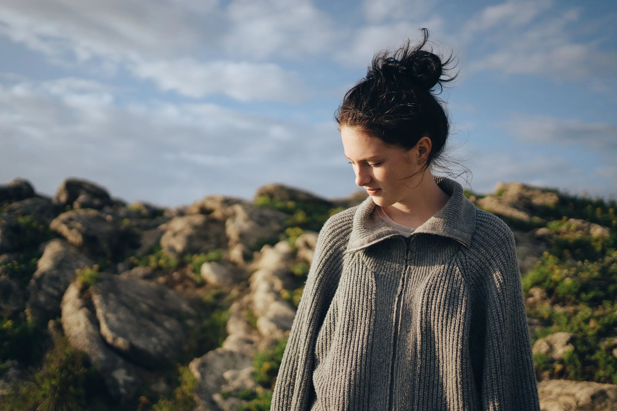 Junge Frau mit braunen, lockigen Haaren in einem Zopf trägt einen grauen Pullover und steht vor einer felsigen Landschaft unter einem bewölkten Himmel.