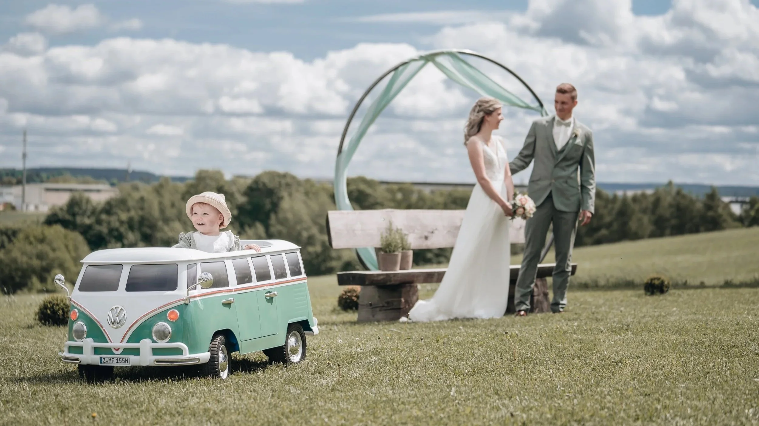 Ein kleines Kind sitzt in einem Spielzeugauto in Form eines VW-Bullis, während ein Brautpaar im Hintergrund sich vor einem dekorierten Holzbalken küsst, auf einer grünen Wiese bei schönem Wetter.