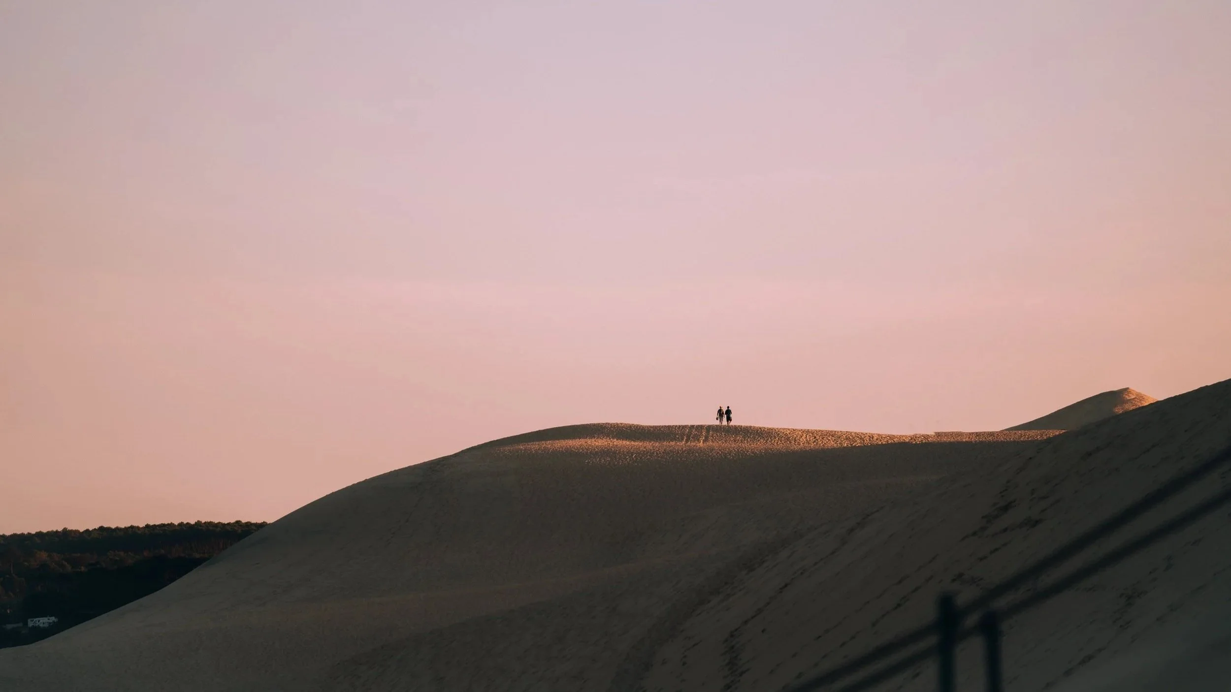 Zwei Personen stehen auf einer Sanddüne bei Sonnenuntergang, im Hintergrund ein sanft erleuchteter Himmel.