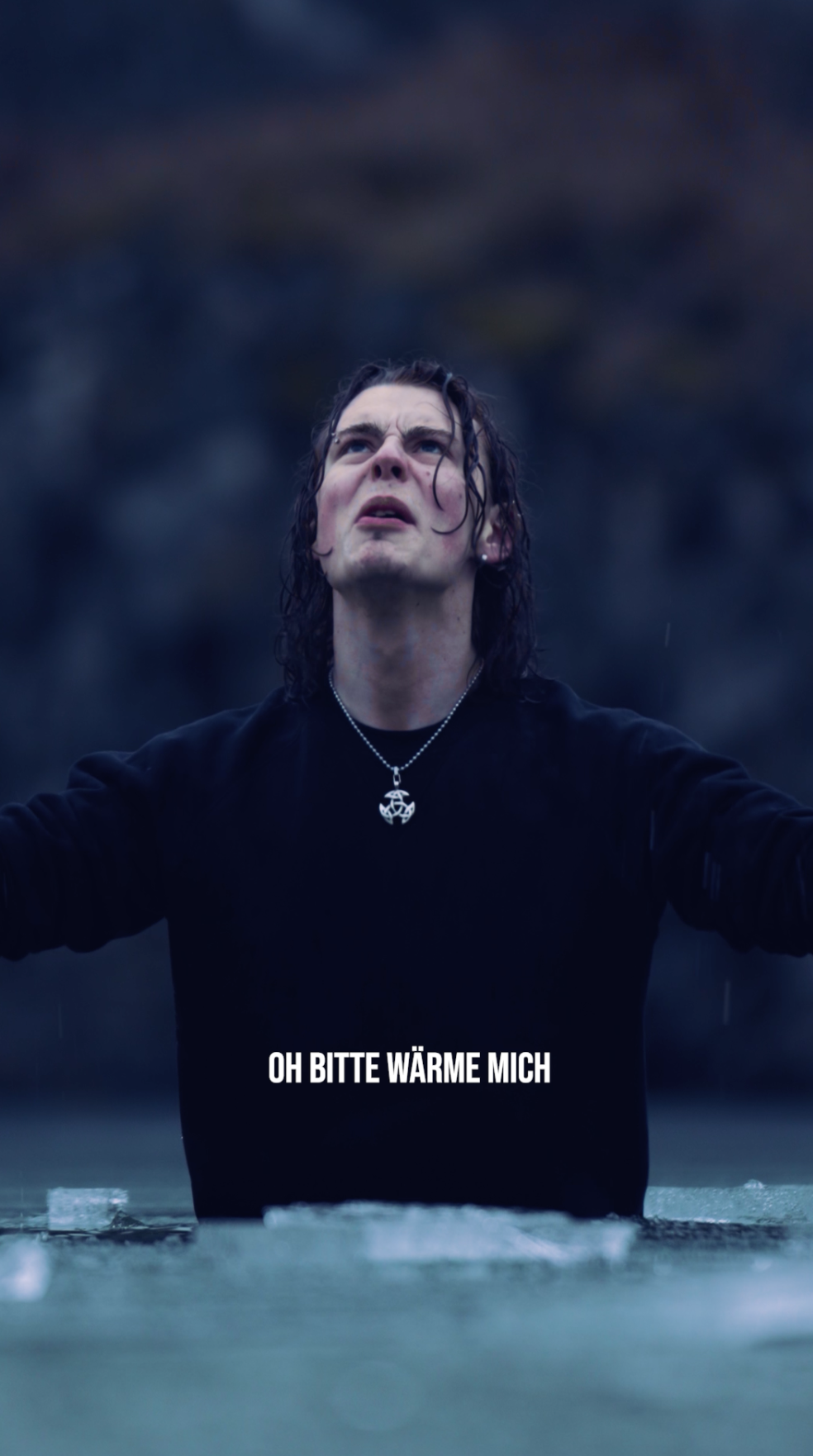 Junger Mann mit nassem langen schwarzen Haaren trägt schwarzen Pullover und Kette mit Anhänger, steht im Wasser mit ausgebreiteten Armen, Blick nach oben, triste Stimmung, Text auf Bild: 'OH BITTE WÄRME MICH'.