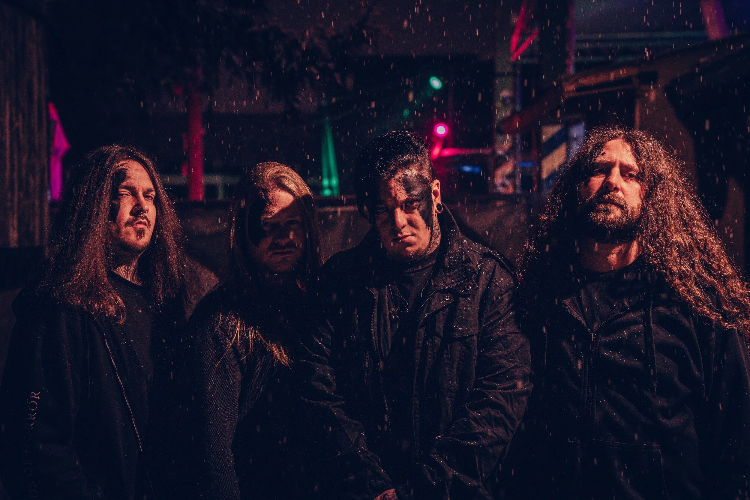 Vier Männer in dunkler Kleidung stehen eng zusammen in einer dunklen, regnerischen Umgebung mit bunten Lichtern im Hintergrund.