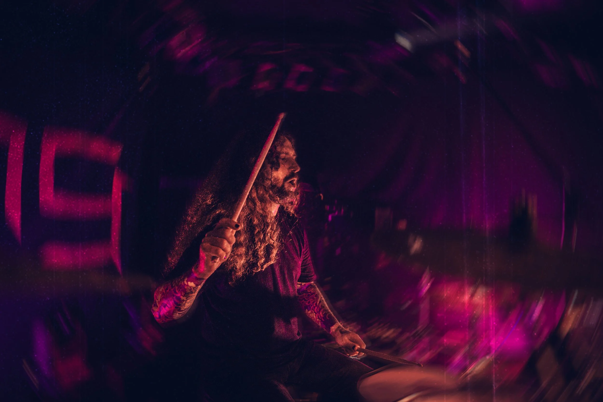 Ein Dynamisches Bild eines Schlagzeugers während eines Auftritts. Der Hintergrund verwirbelt sich um ihn. Er holt zum Schlag aus. Das Licht ist leuchtend Violet.