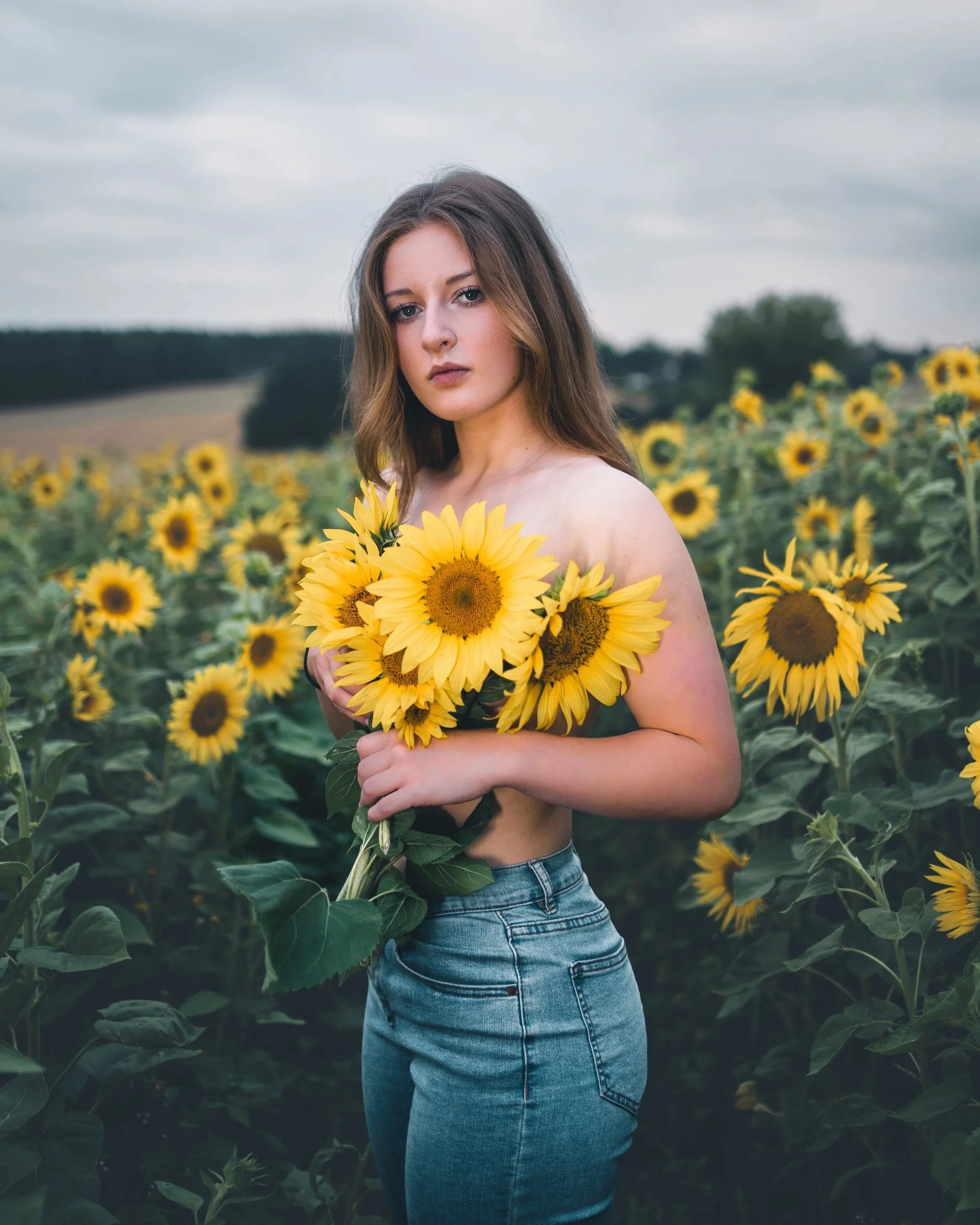 Junge Frau in Jeans, die in einem Sonnenblumenfeld steht und Sonnenblumen hält.