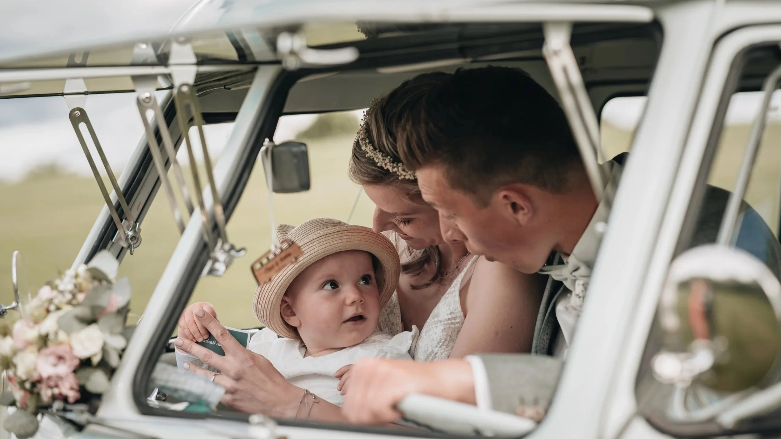 Familie in einem Oldtimer, das Baby trägt einen Strohhut, die Eltern umarmen das Kind.