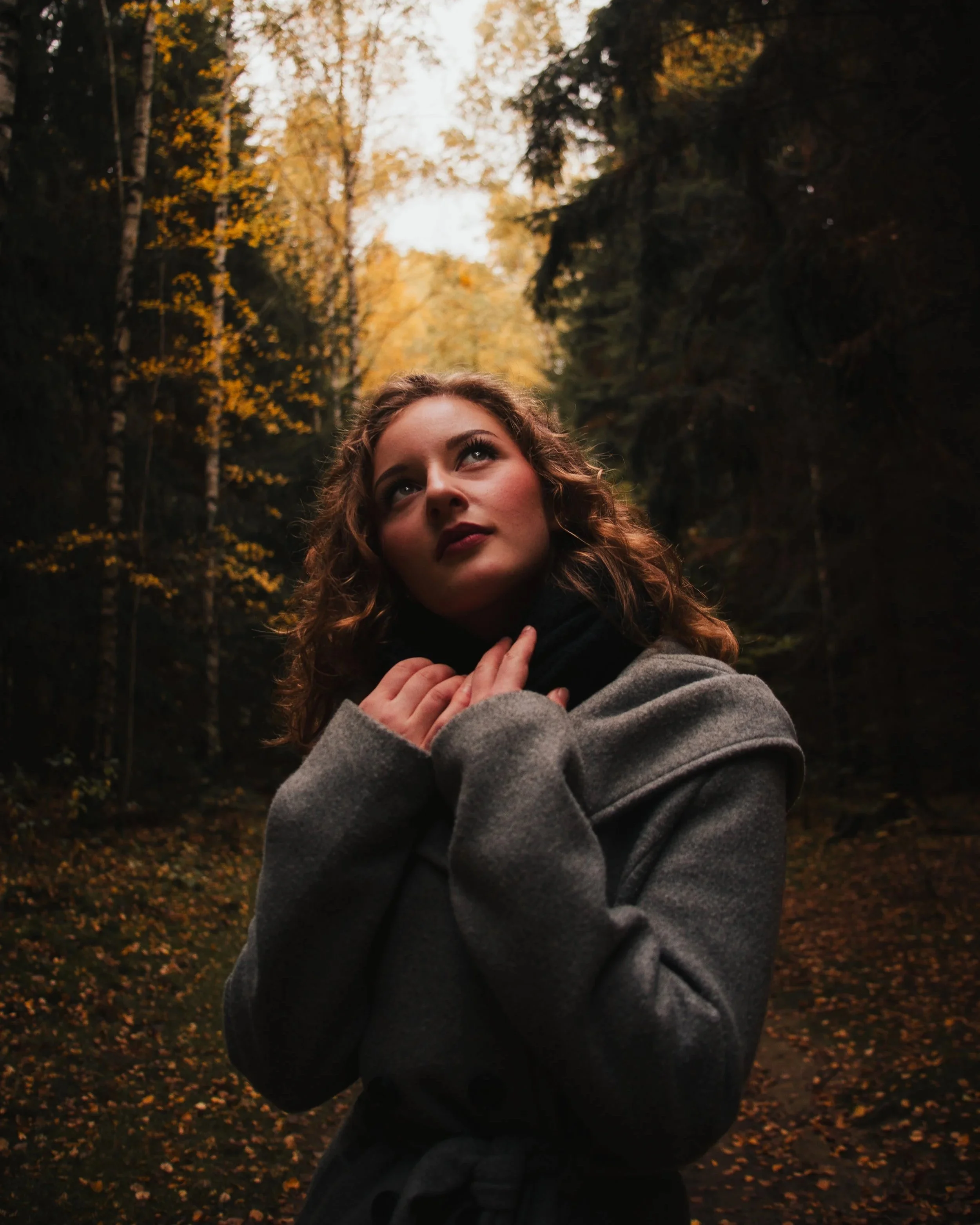 Eine junge Frau mit lockigen Haaren und dunklem Lippenstift steht in einem herbstlichen Wald, trägt einen grauen Mantel und blickt nach oben.