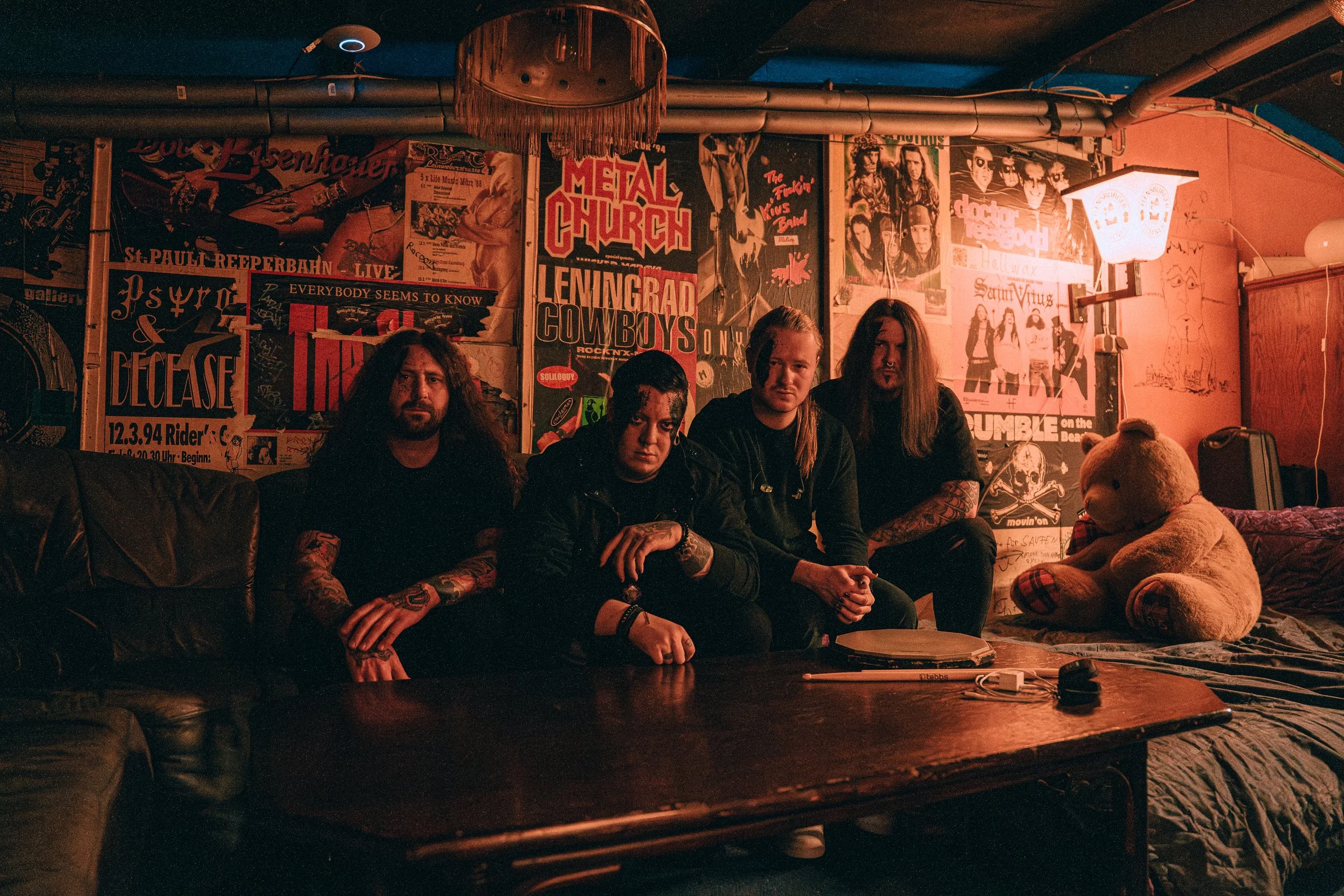 Eine Band bestehend aus vier Personen. Schwarz geschminkt. Der Hintergrund ist mit Plakaten verkleidet. Rechts ein großer Stoffbär und eine Laterne. Die Musiker sitzen auf einer Couch.