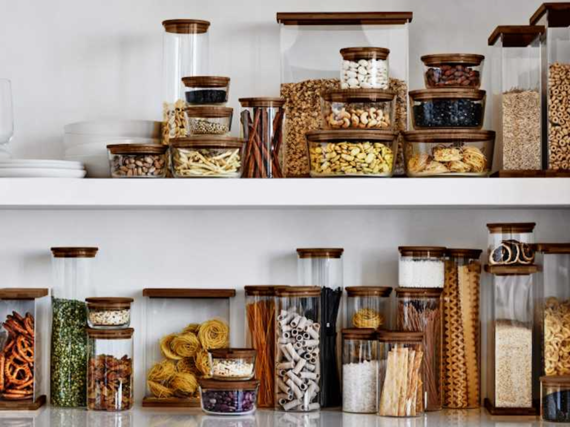 A kitchen shelf holding multiple glass jars filled with pasta, spices, and other dry ingredients, with some jars topped with wooden lids.