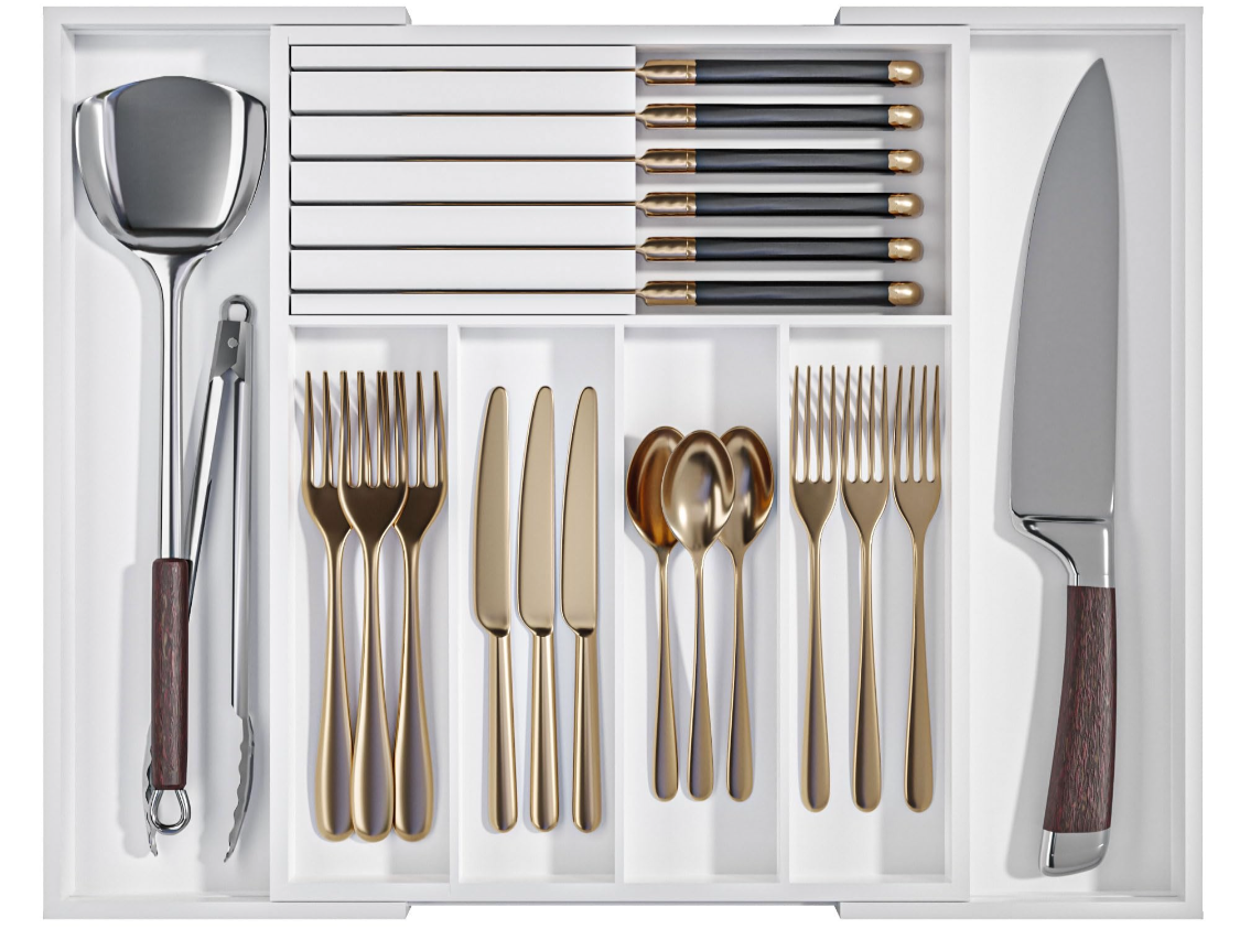 A neatly organized set of kitchen utensils including a large slotted spoon, a box of six steak knives, six black-handled butter knives, six golden forks, six golden teaspoons, six golden knives, and six golden dinner forks, all stored in a white cutlery drawer.