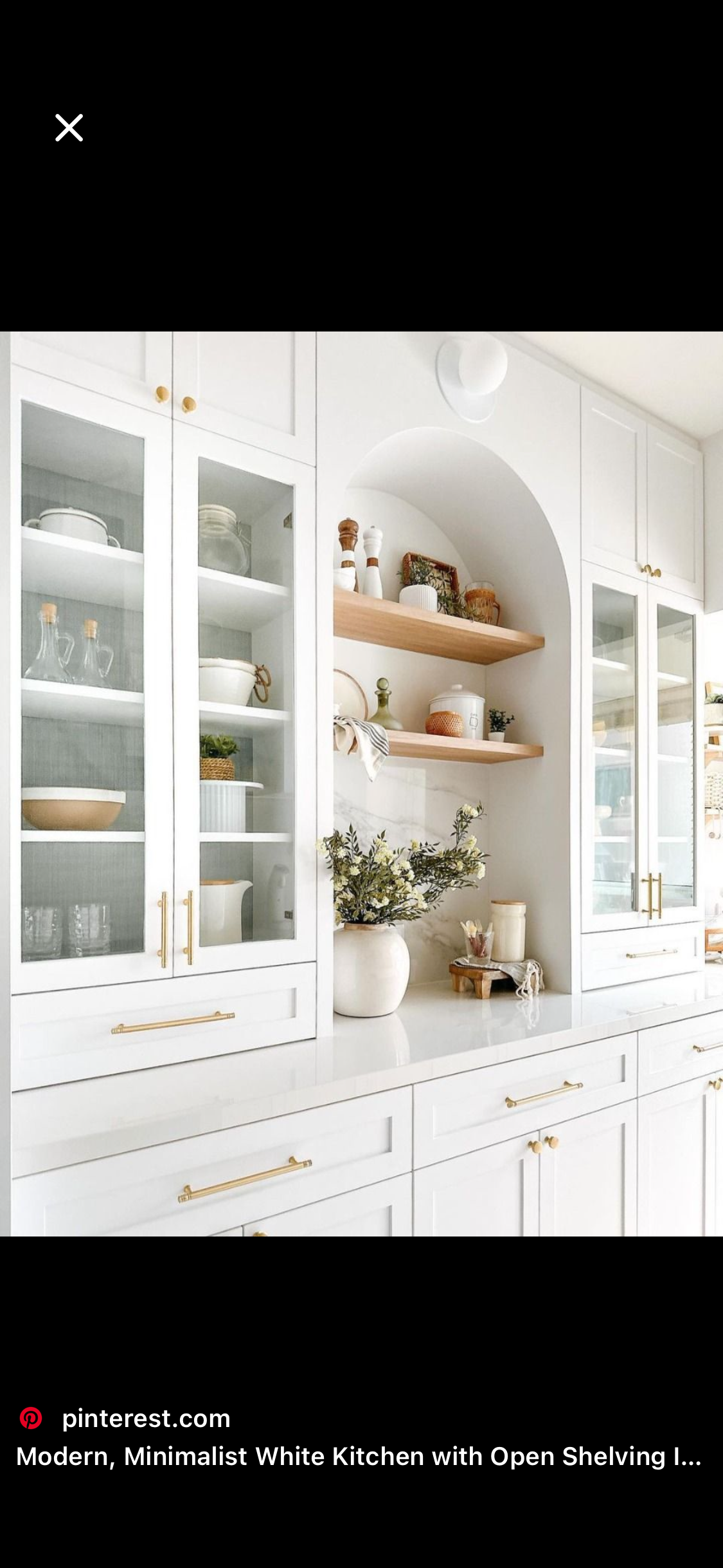 A modern white kitchen with glass-front cabinets, open wooden shelves, and gold accents, decorated with plants and kitchenware.