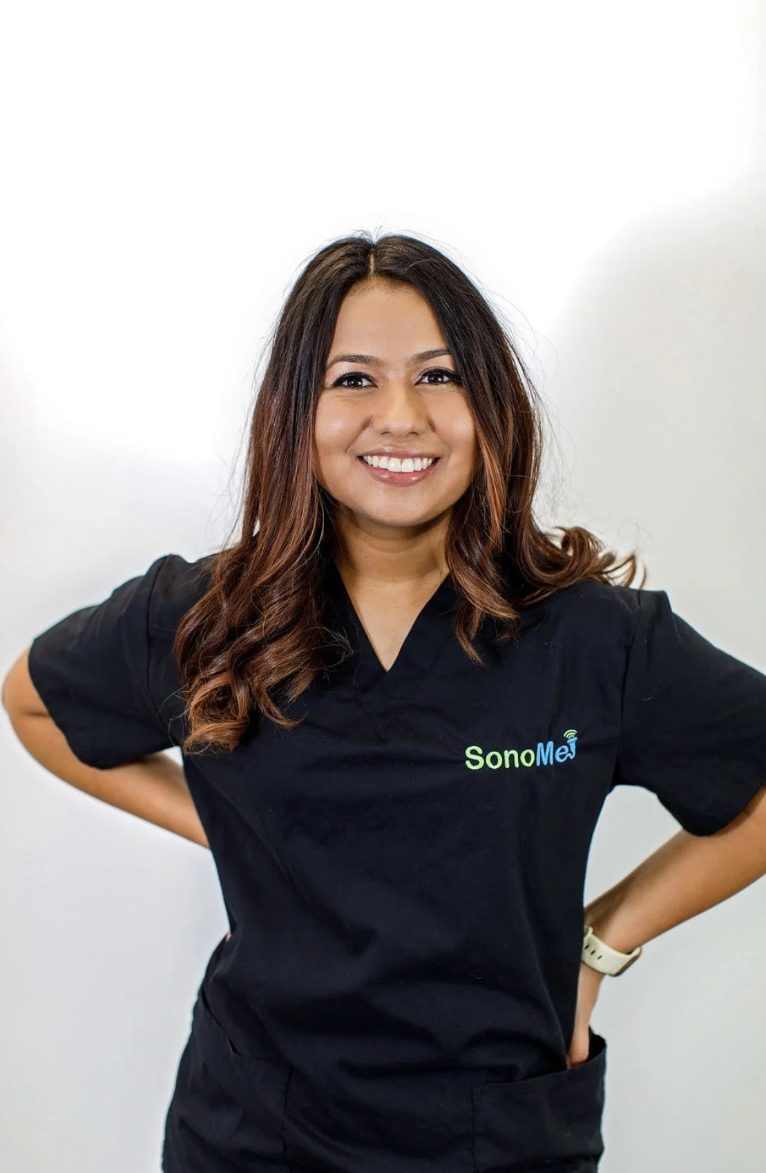 A woman smiling and posing with her hands on her hips, wearing black scrubs with the logo 'SonoMe' on it.