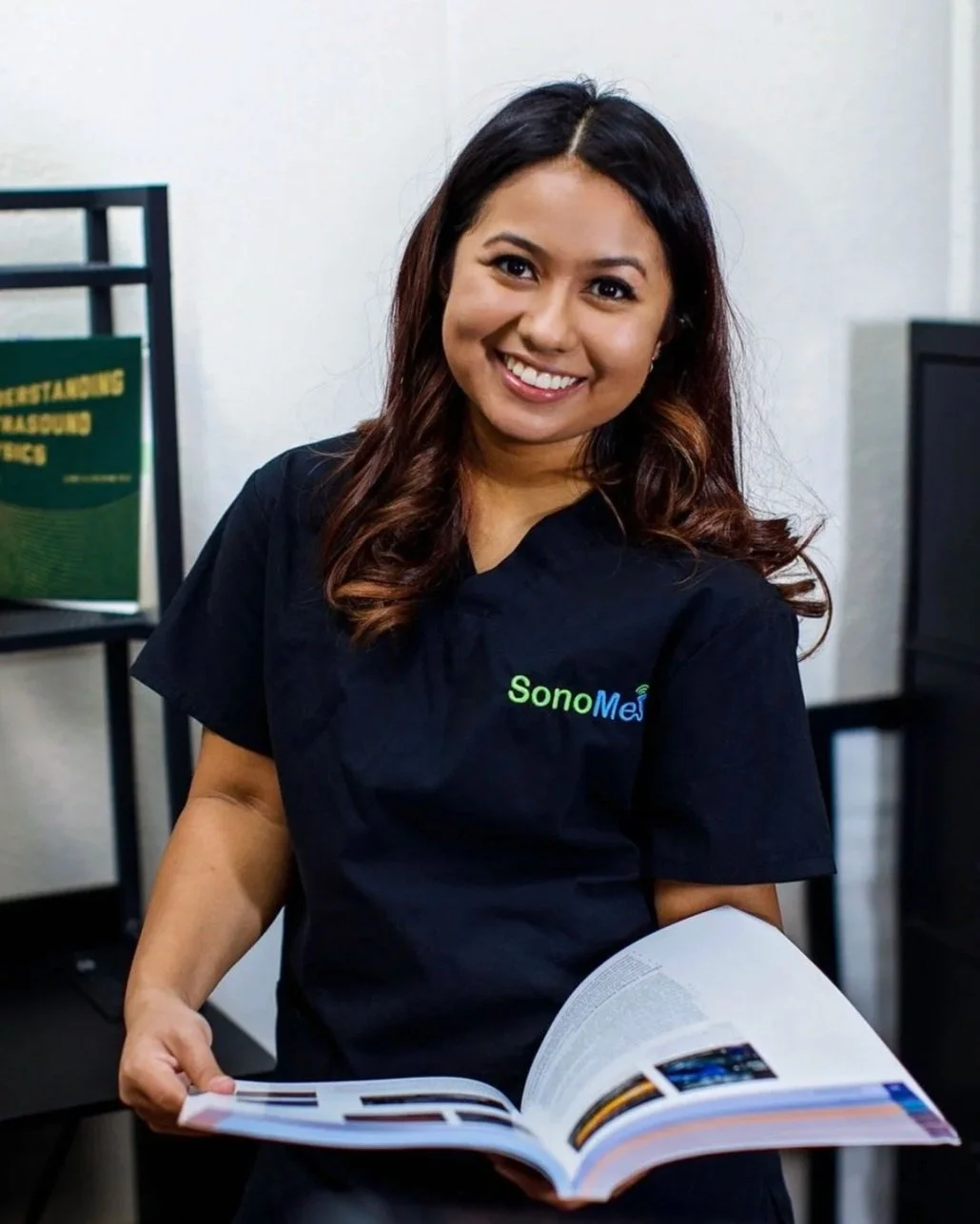 A woman smiling and holding an open book in an office setting. She is wearing a black shirt with a logo that says 'SonoMe.'