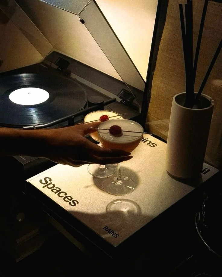 A hand holding two cocktails with cherries on a table near a vintage record player and a plant in a white pot.