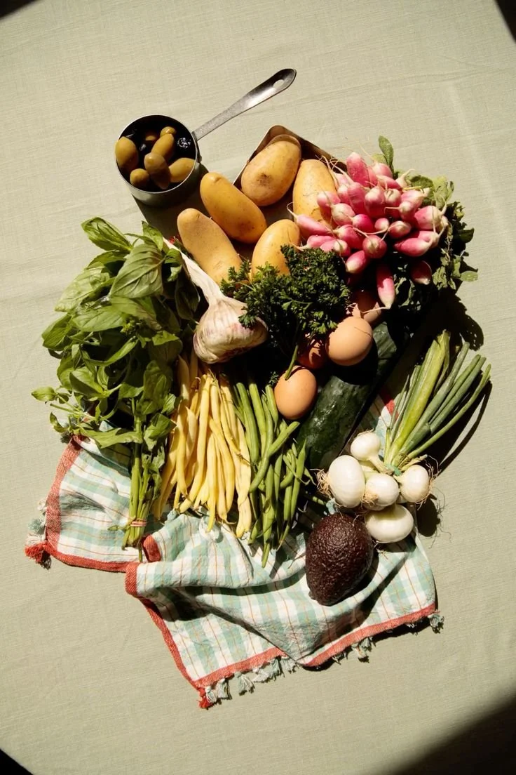 A variety of fresh vegetables and eggs arranged on a checkered cloth, including radishes, potatoes, cucumbers, garlic, spring onions, green beans, eggs, basil, and an avocado, with a small can of olives.