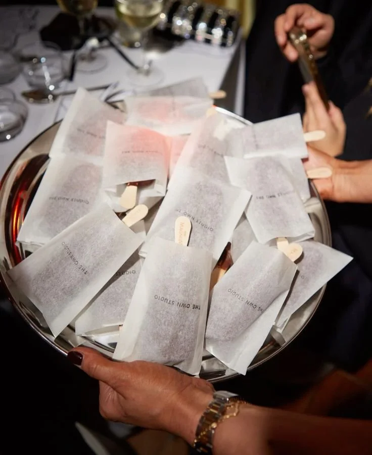 A person holding a tray with multiple wrapped mints or candies, with a wine glass and other tableware in the background.