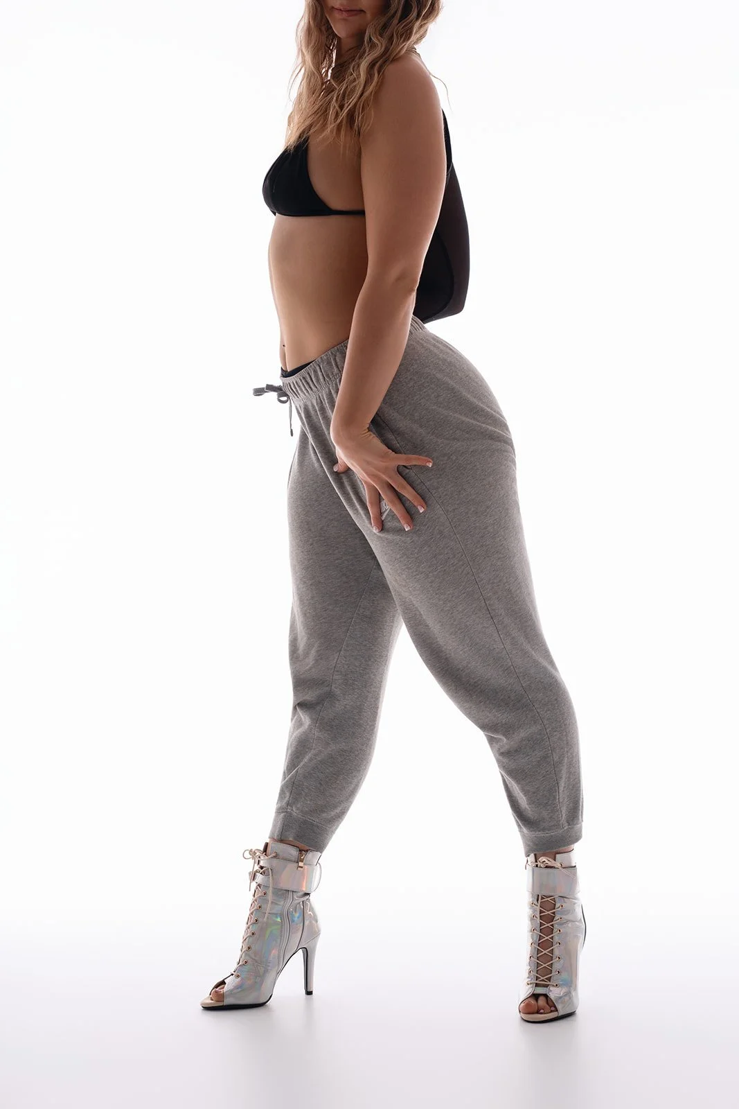A woman with wavy blonde hair wearing a black sports bra, gray sweatpants, and high-heeled iridescent boots, standing against a white background