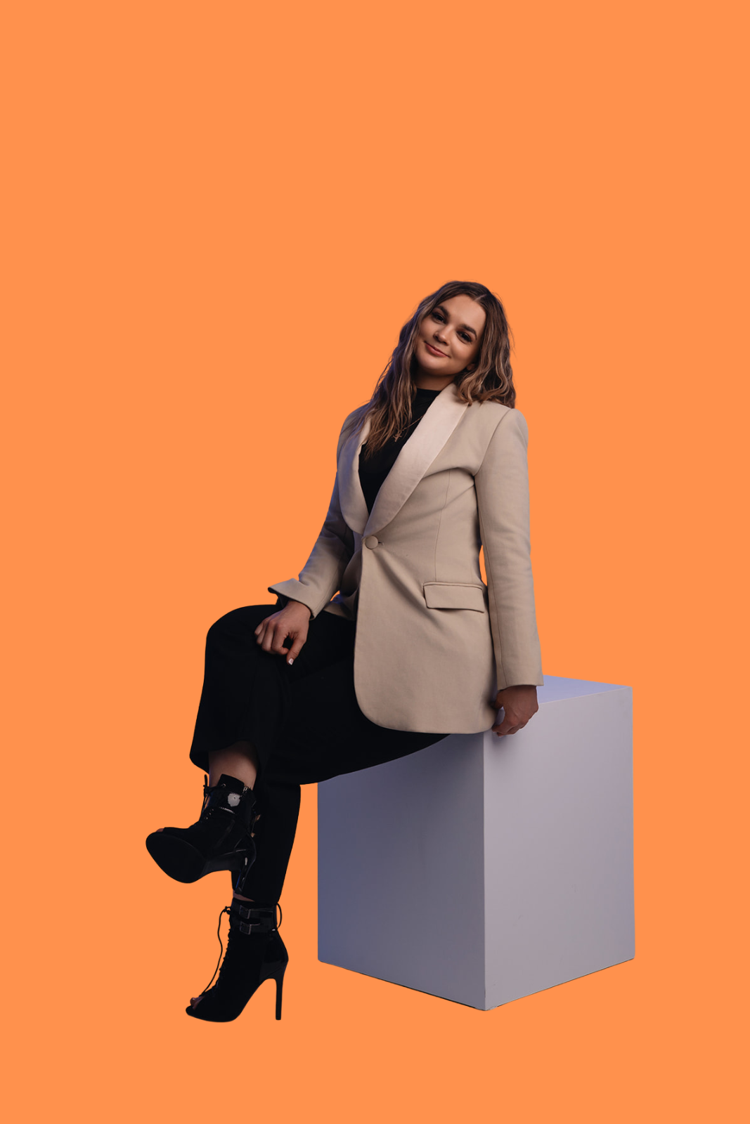 A woman with wavy hair sitting on a white block against an orange background, wearing a beige blazer, black dress, and high-heeled boots.