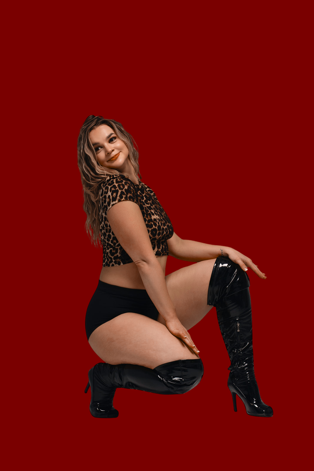 Woman with brown hair in a leopard print crop top and black shorts, kneeling on one knee against a red background, wearing shiny black thigh-high boots.