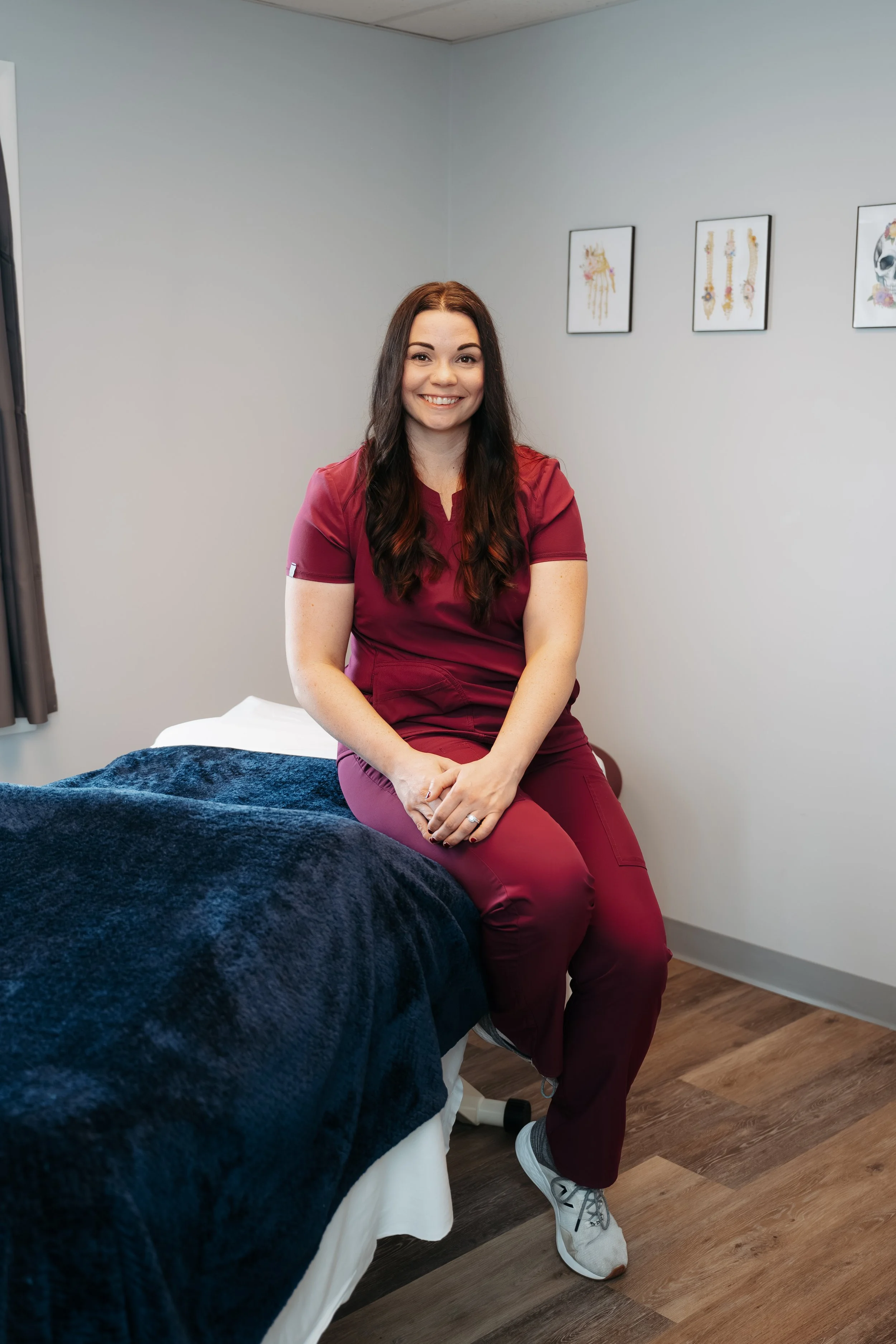 A woman in maroon scrubs sitting on the edge of a treatment bed in a medical office, smiling at the camera.