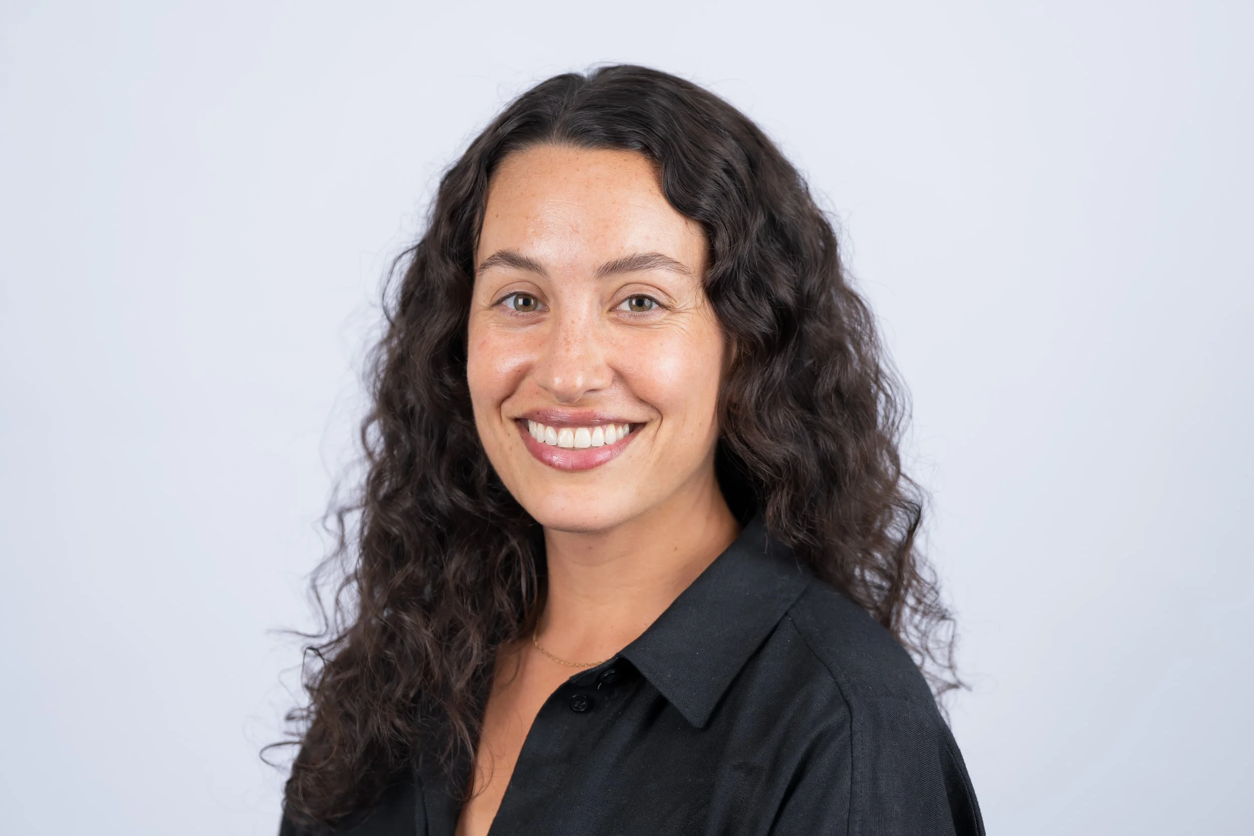 A woman with long, curly dark hair, wearing a black top, smiling, against a plain light gray background.