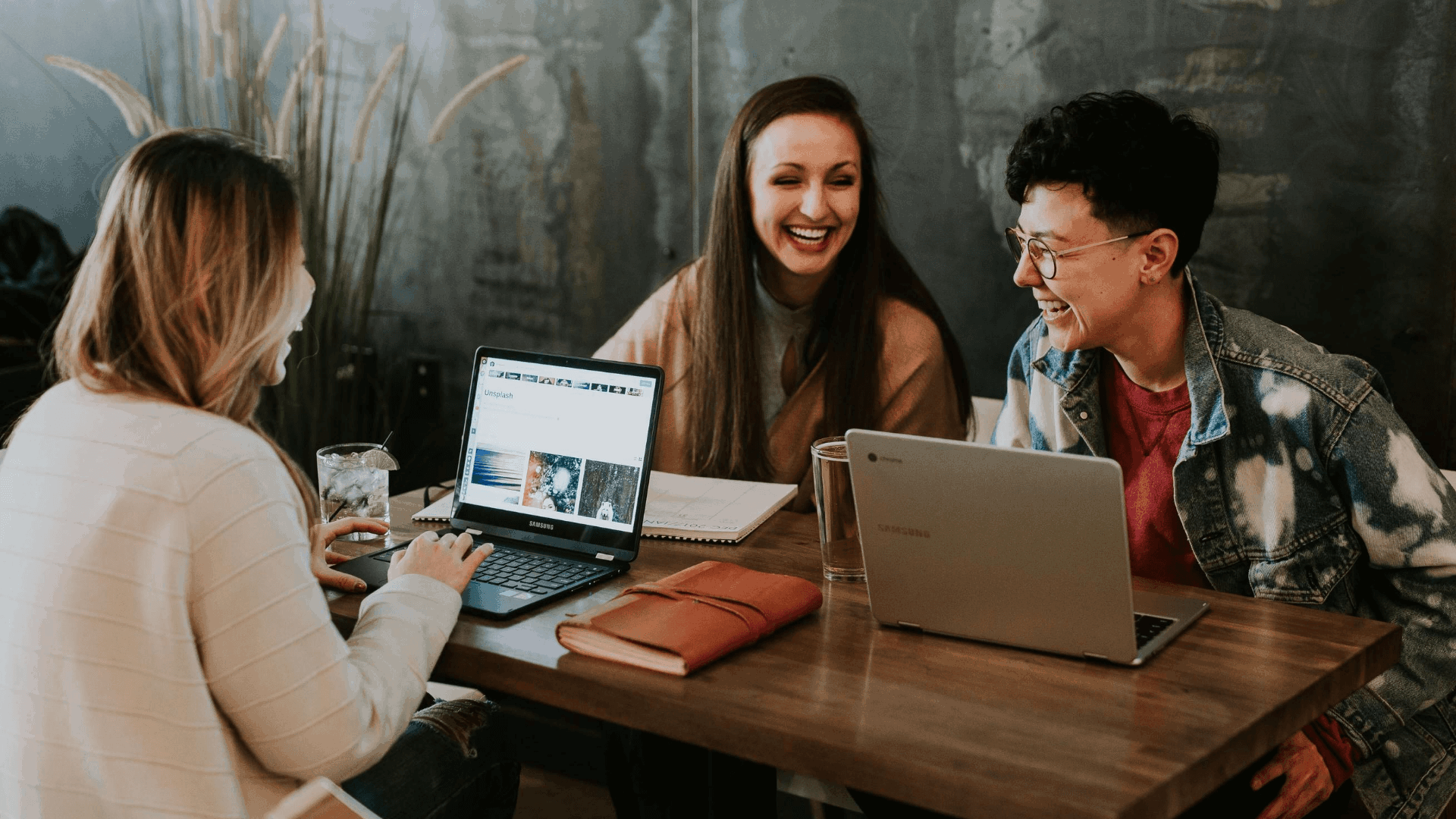 Three friends are sitting at a wooden table in a cafe, laughing and chatting. Two laptops and a tablet are on the table, along with a glass of water and a notebook. The background shows a dark, textured wall and some plants.