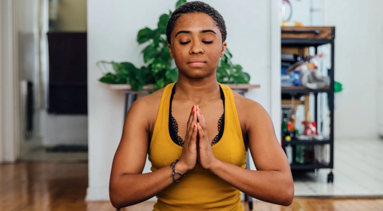 Young woman practicing meditation at home with hands in prayer position, eyes closed, sitting cross-legged.