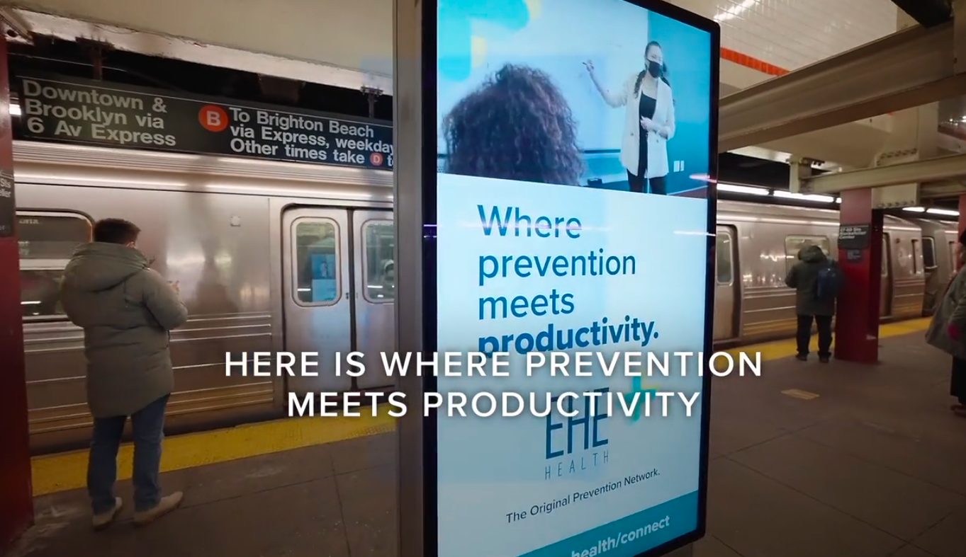 Subway station platform with a digital advertisement display reading "Where prevention meets productivity" and showing a healthcare professional wearing a mask. There are a few people waiting, and a subway train is in the background.