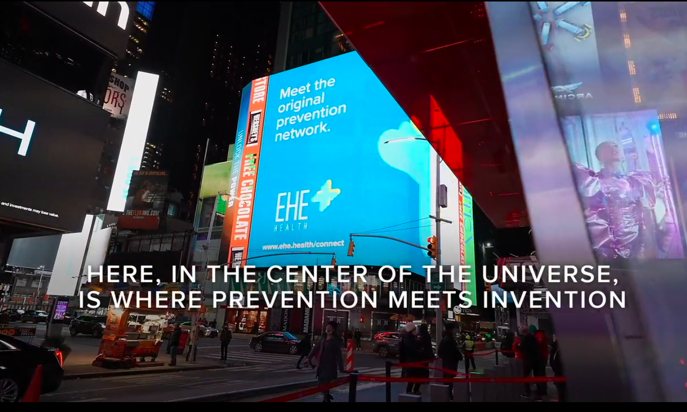 A busy city street scene at night with large digital billboards including one advertising a health prevention network, and people crossing the street under traffic lights. Overlaid text reads: "HERE, IN THE CENTER OF THE UNIVERSE, IS WHERE PREVENTION MEETS INVENTION."