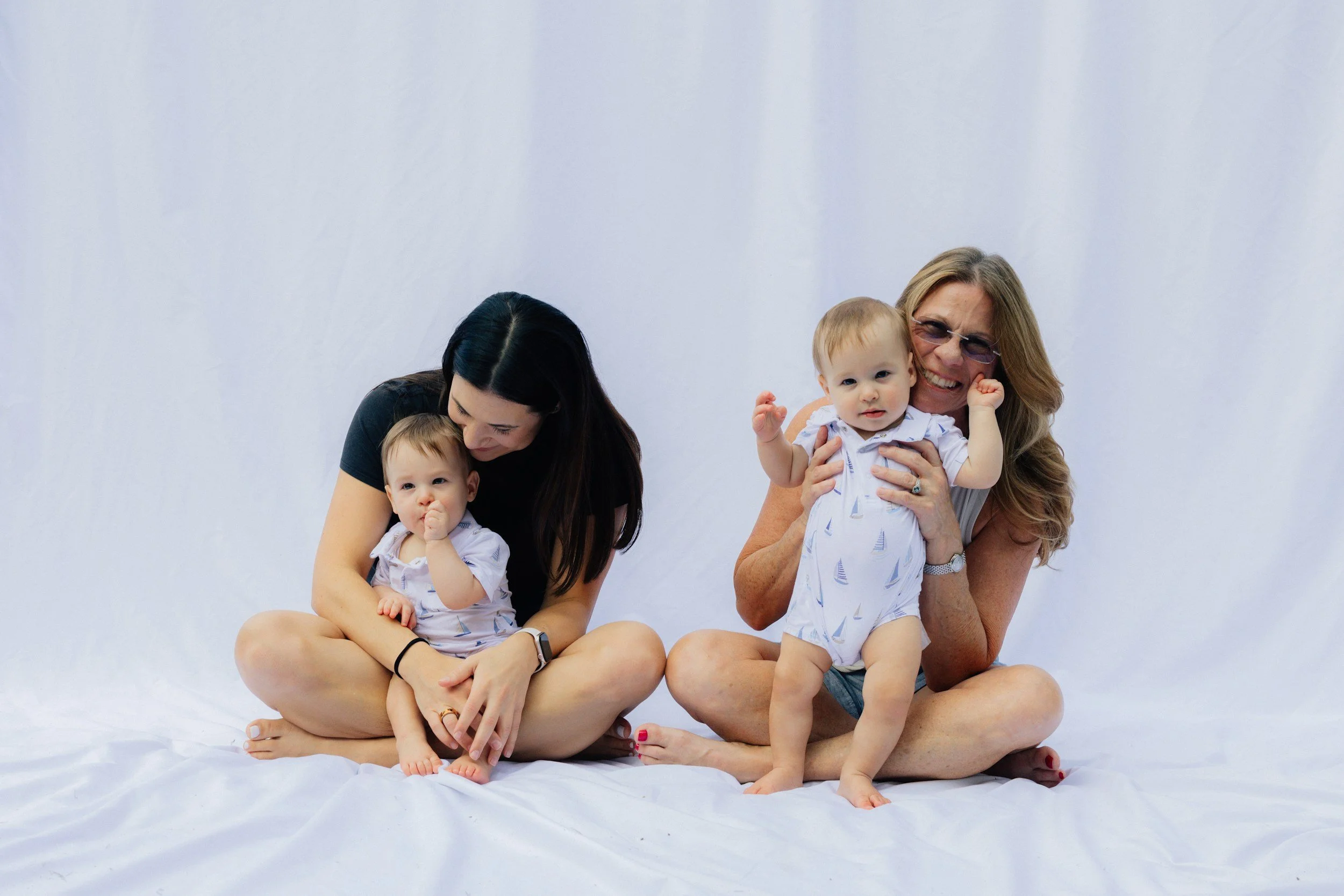Two women, one with light skin and long brown hair and the other with light skin and long blonde hair, sitting on the floor with two young children, all wearing matching white outfits and smiling against a plain white background.