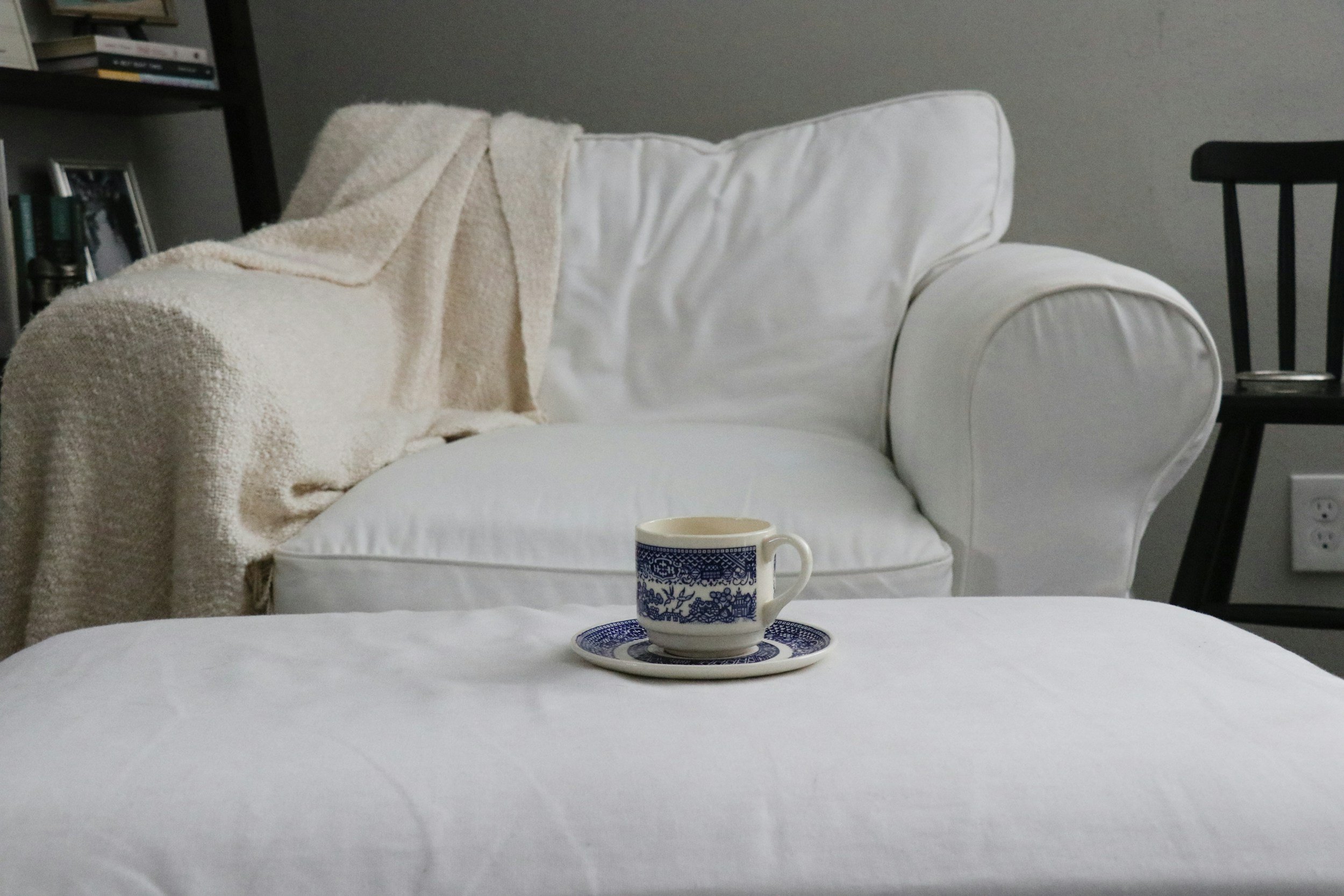 A white table with a blue and white patterned coffee cup on a saucer, in front of a white sofa with a beige blanket draped over the armrest, and a bookshelf and black chair in the background.