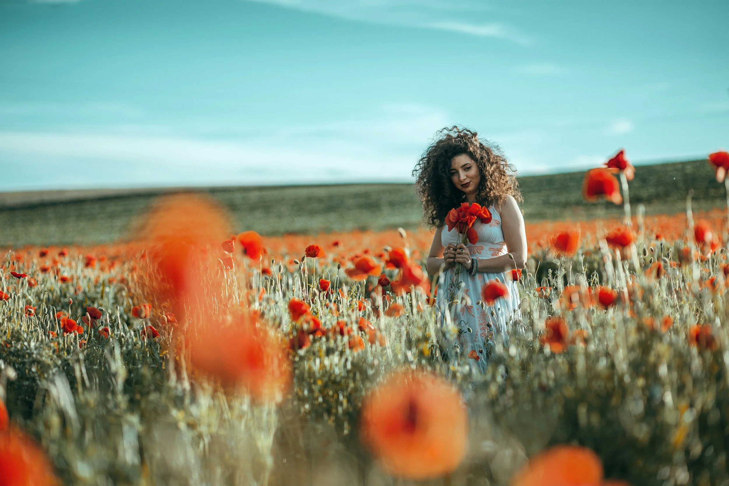 A woman with curly hair in a white dress stands in a field of red poppies, holding a bouquet of flowers, with a hilly landscape and blue sky in the background.