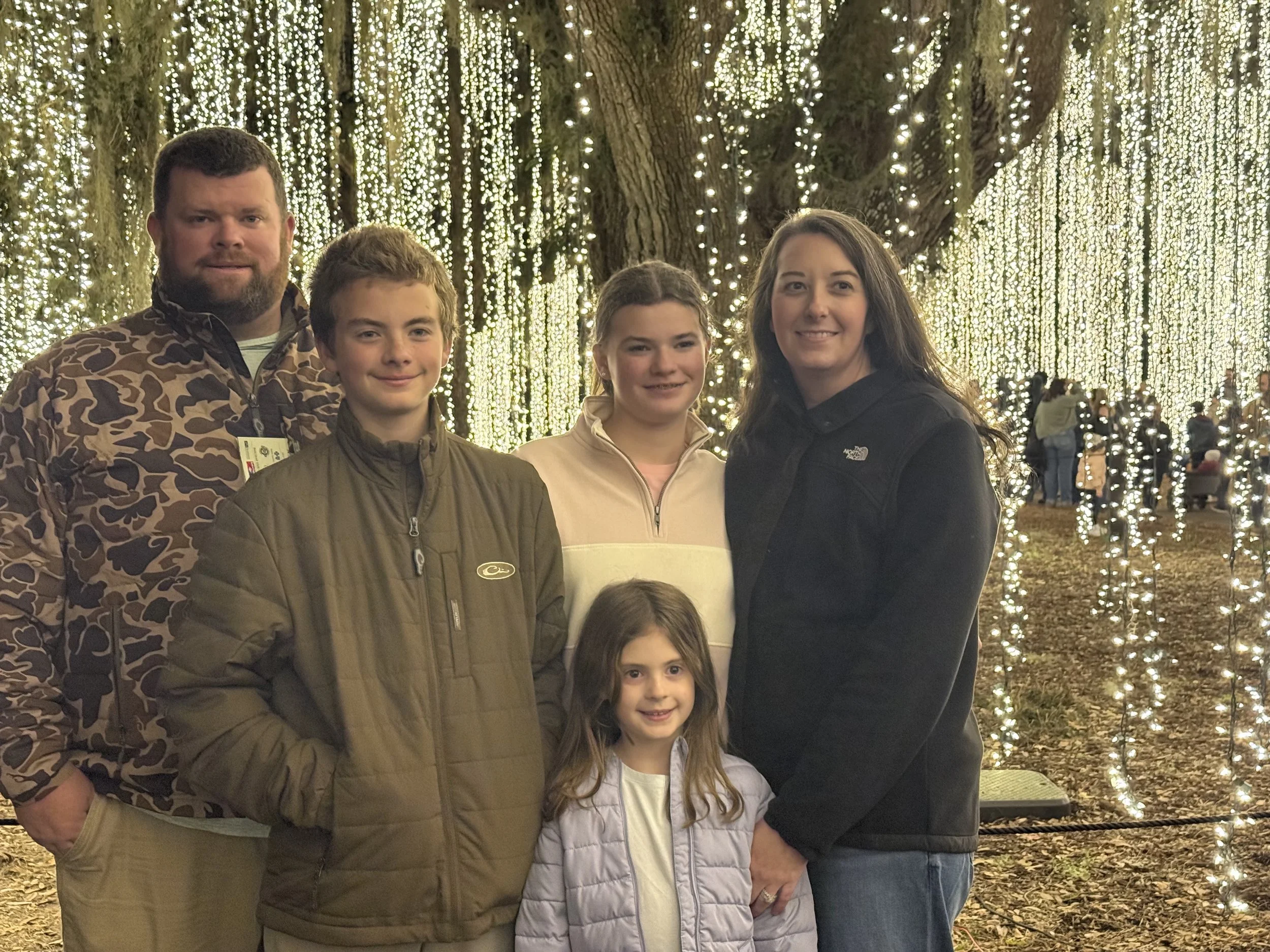 A family of five standing in front of a backdrop of Christmas lights on a tree, with other people in the background, during an outdoor winter event.