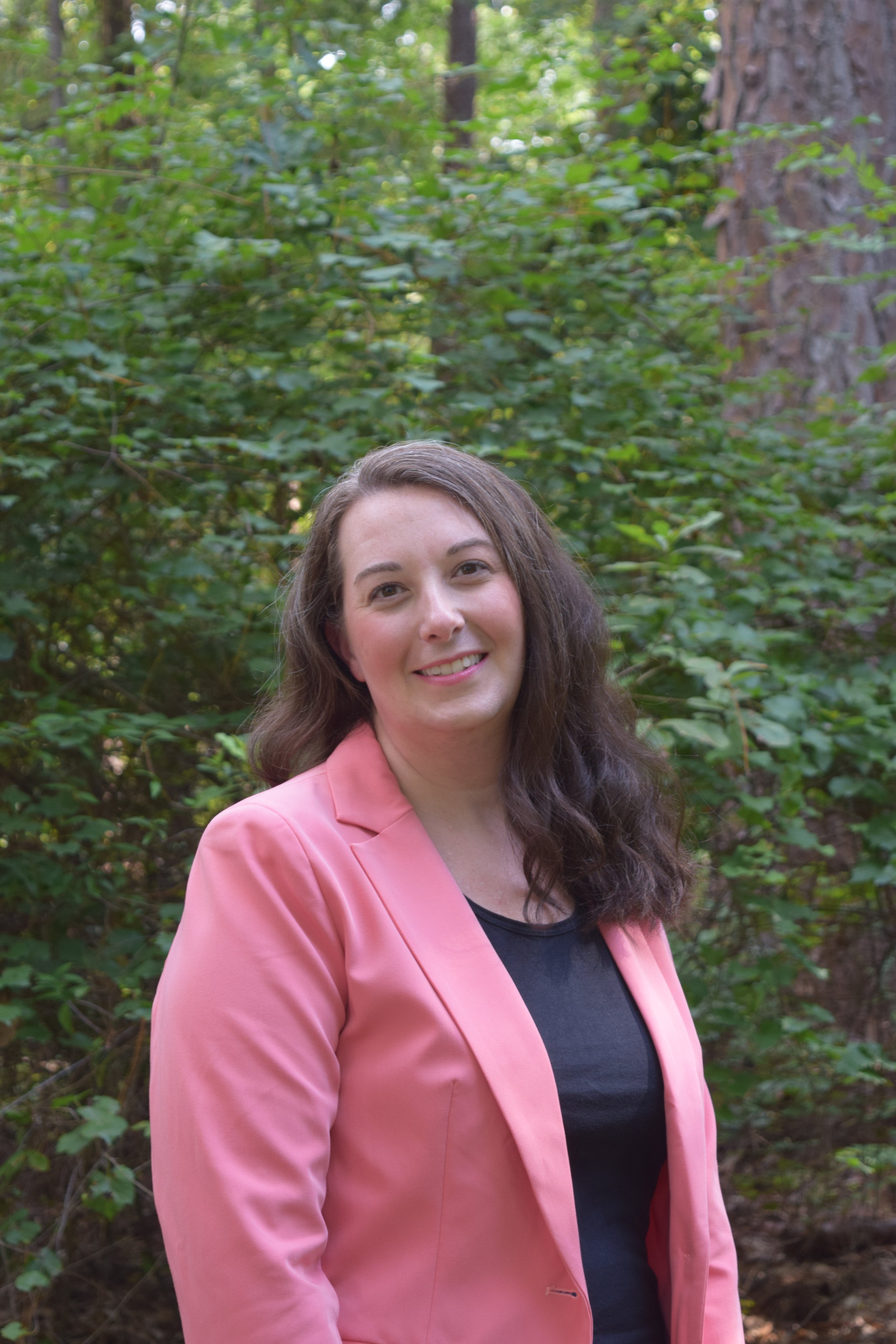 A woman with brown, wavy hair wearing a pink blazer and black top, standing outdoors in front of green foliage and trees, smiling at the camera.