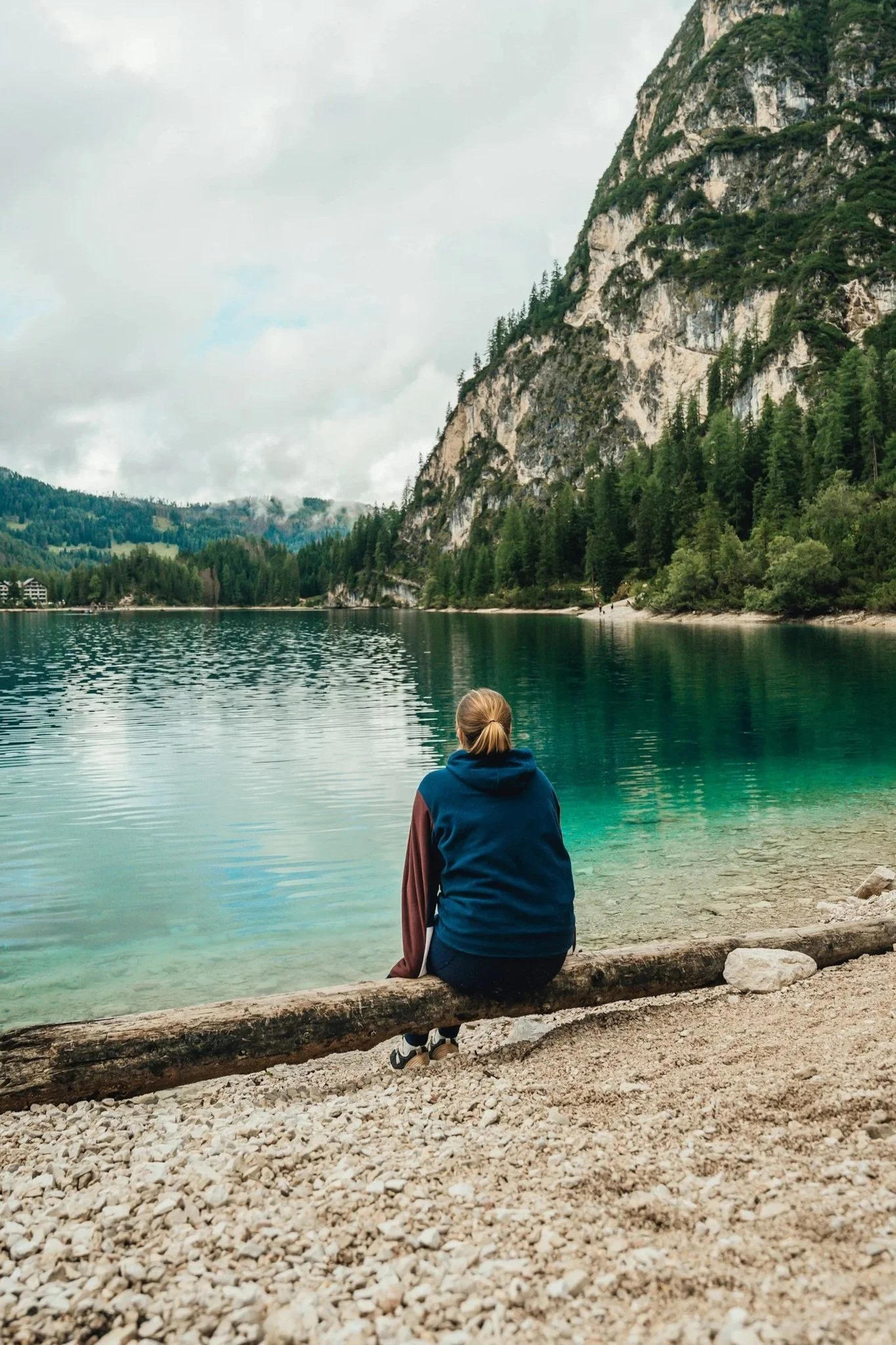 A person with blonde hair sitting on a log by a calm, green lake, surrounded by tall, forested mountains under a cloudy sky.