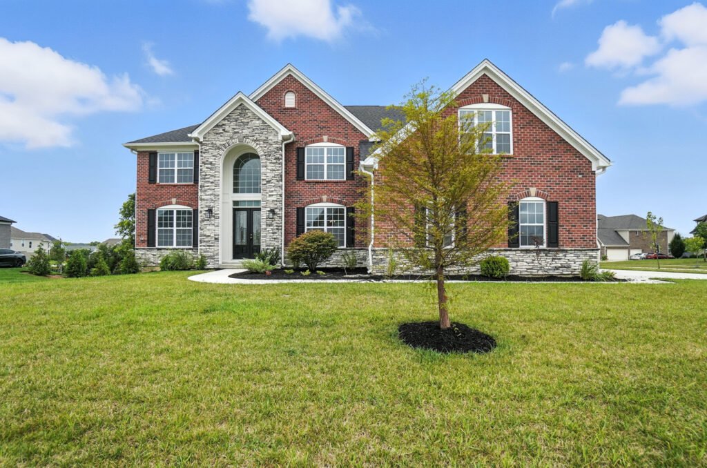 A large two-story brick house with a stone facade, multiple windows, and a well-maintained lawn with a small tree in the front yard.