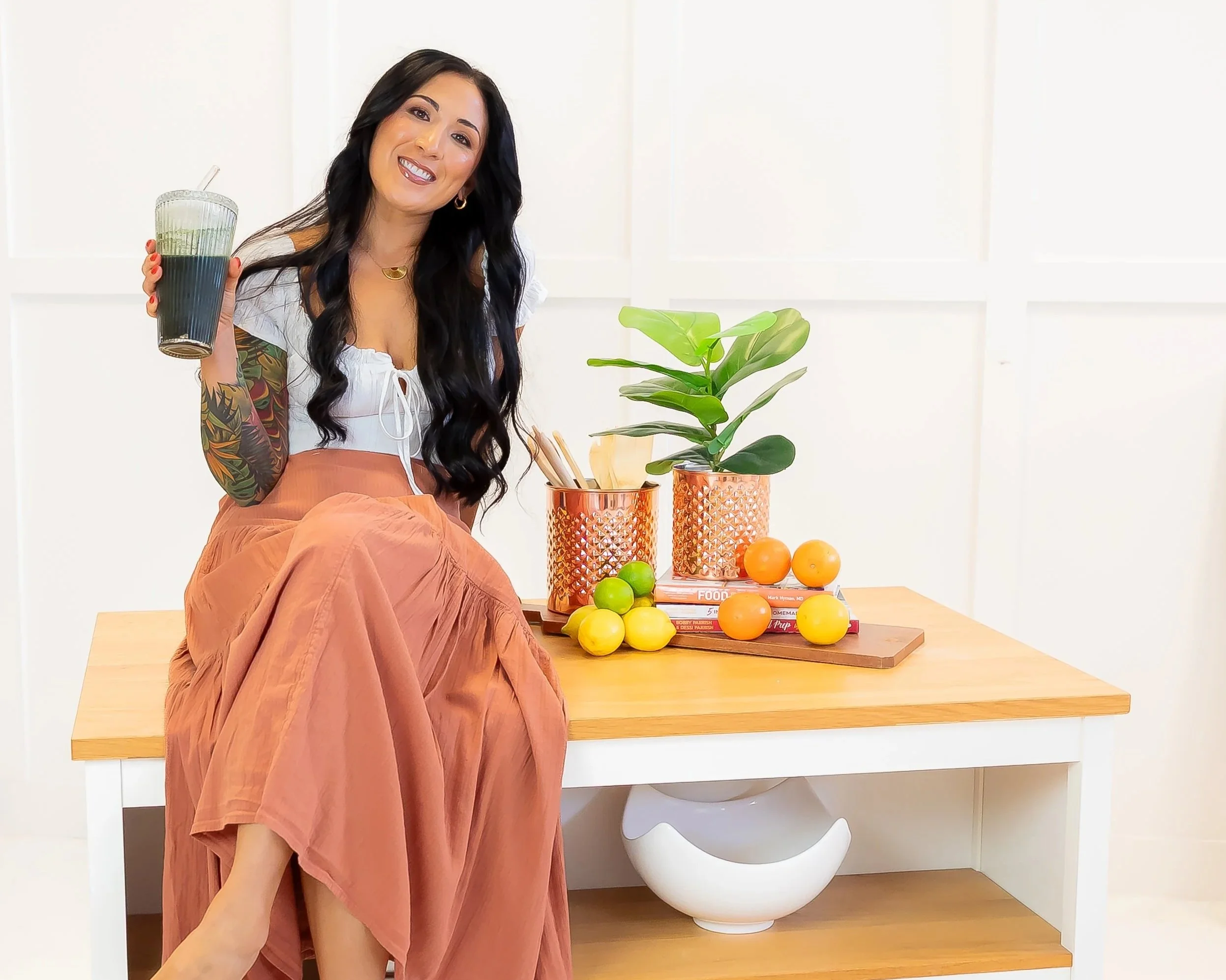 A woman sitting on a wooden table, holding a glass of dark-colored beverage, smiling, with a tattoo sleeve on her left arm, wearing a white top and a peach-colored skirt, with a white and orange decorated background, and small trees and fruits on the table.