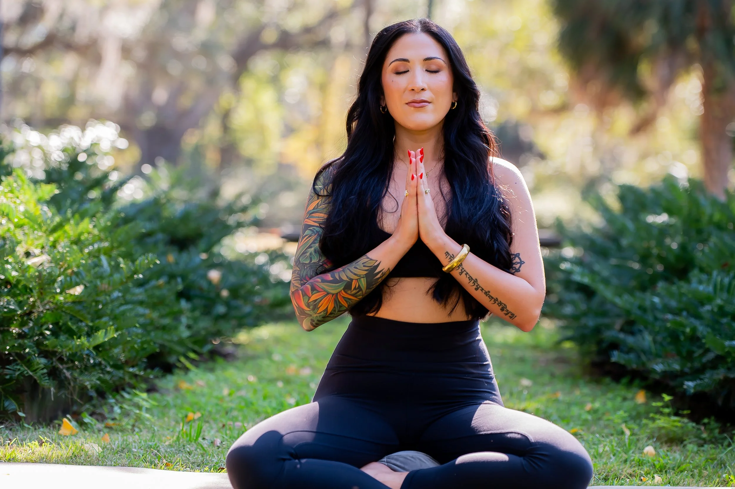 A woman with long dark hair and colorful tattoos on her left arm practicing yoga outdoors, sitting cross-legged with hands in prayer position, eyes closed, in a peaceful setting with greenery and trees in the background.