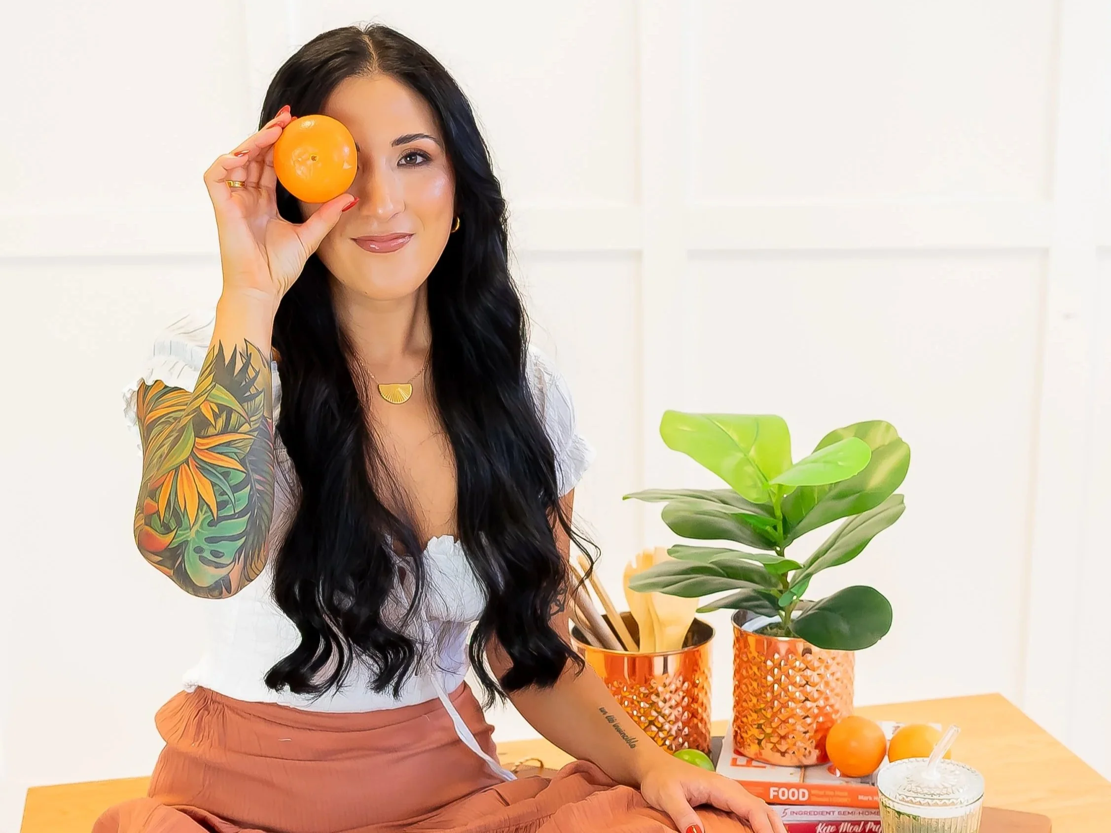 A woman with long black hair, a colorful tattoo sleeve, and a white top, holding an orange fruit to her eye, smiling in a bright room with a plant, books, and a drink on a wooden table.