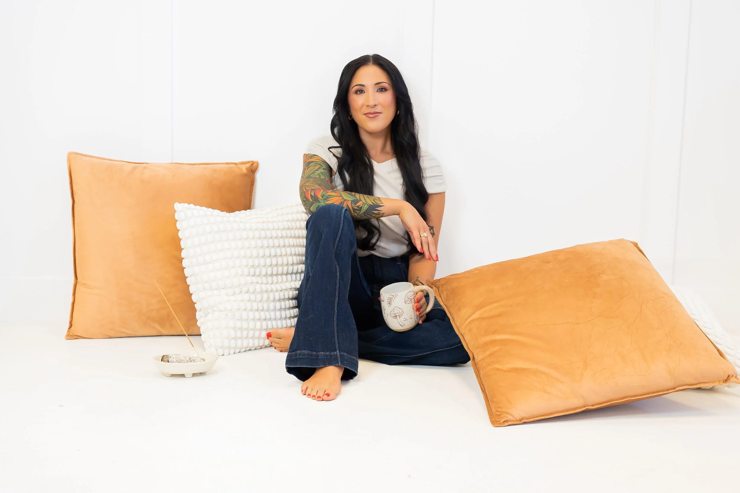 A woman with black hair, tattoos on her arm, sitting barefoot on a white surface surrounded by large pillows in beige and white, holding a mug, with an ashtray nearby.