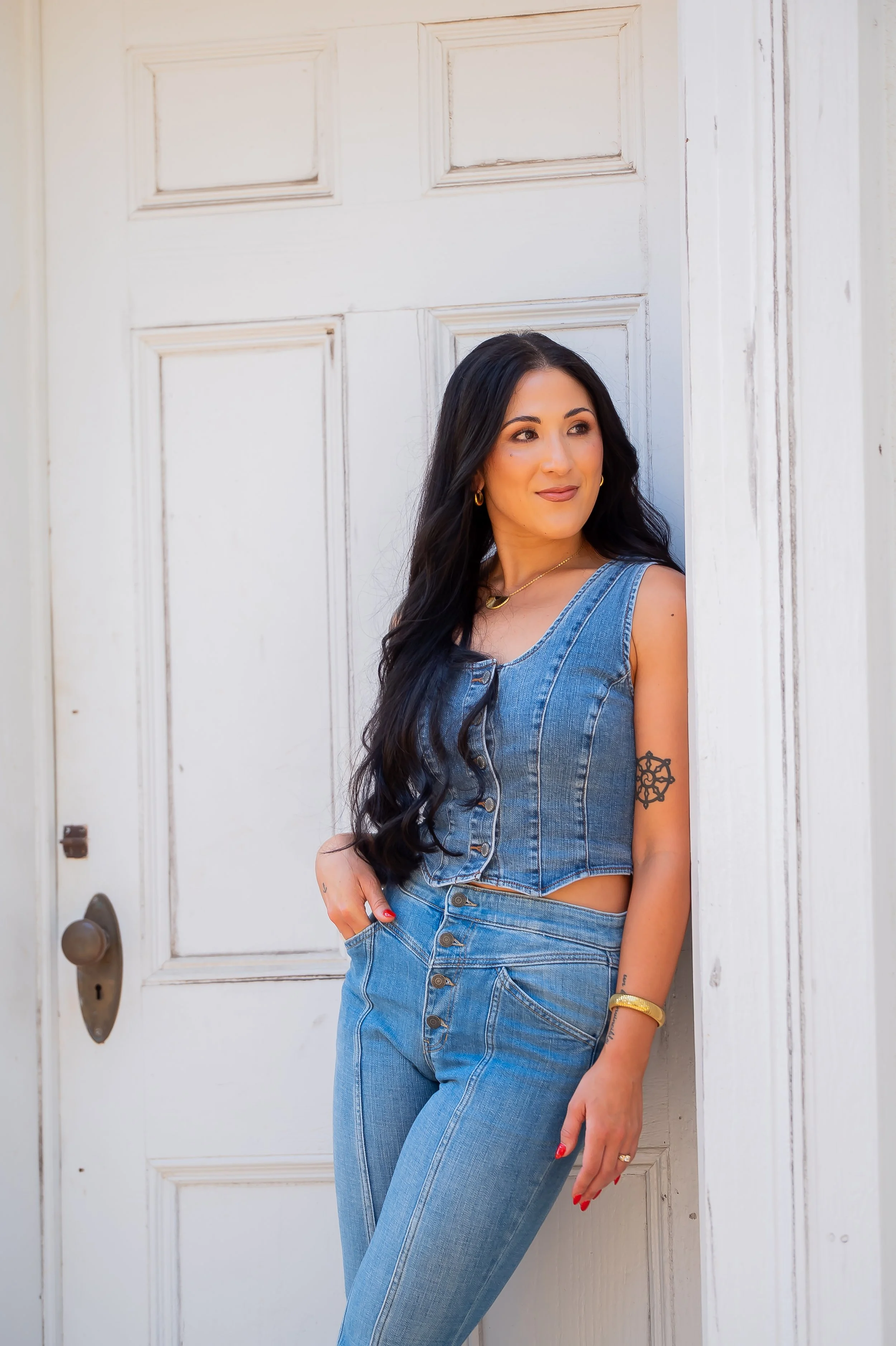 Women with long black hair wearing a sleeveless denim top and jeans, standing in front of a white door, looking to the side.