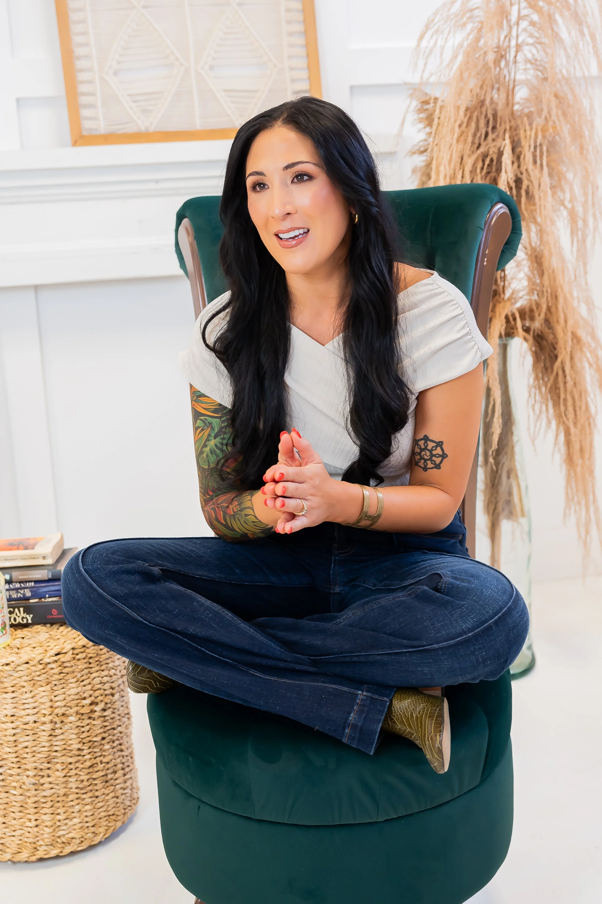 A woman with long black hair, sitting cross-legged on a green chair, wearing a white top and jeans, engaged in conversation in a bright room with decorative dried plants and books.