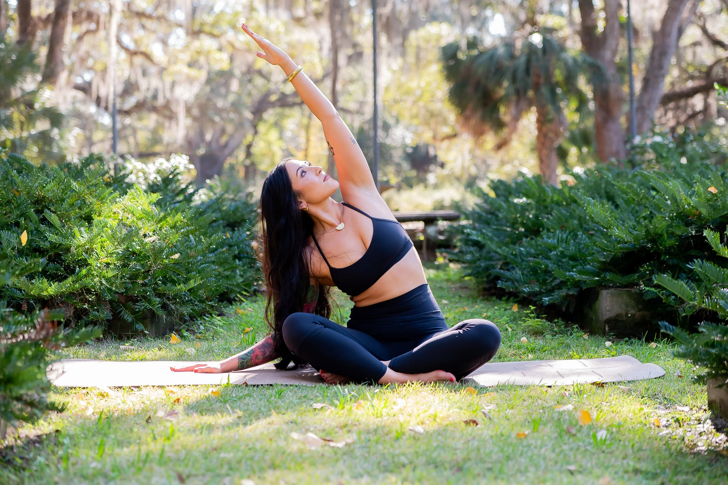 A woman practicing yoga outdoors on a white mat amidst green bushes and trees, wearing black athletic wear, with her hair down, extending her right arm upward while seated in a cross-legged position.