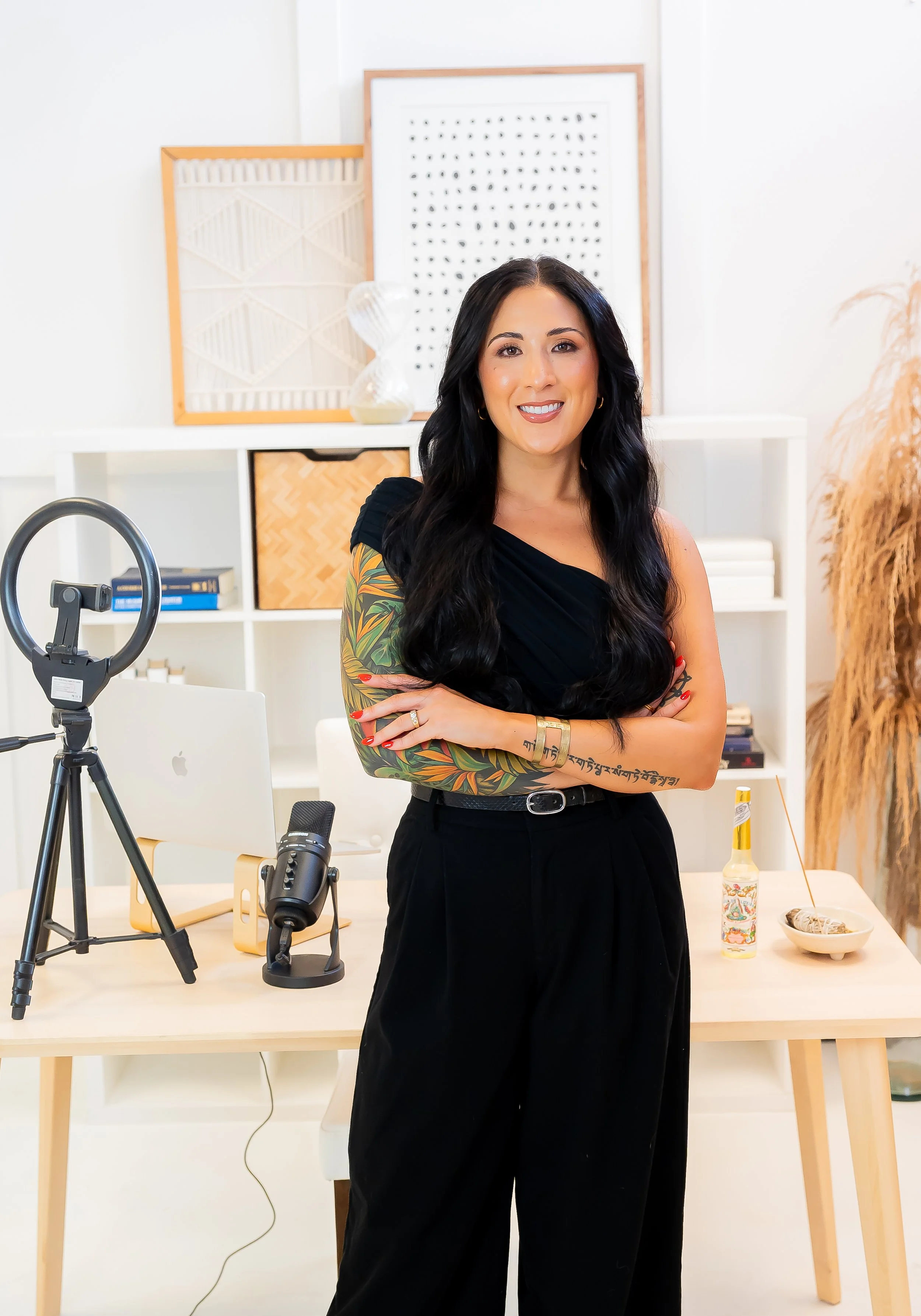 Woman with long dark hair, wearing black, tattoos on her left arm, standing with arms crossed in a modern home office.