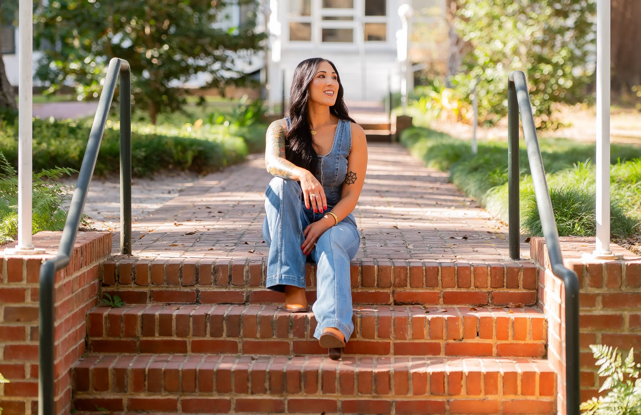 A woman with long dark hair, tattoos, wearing a sleeveless denim jumpsuit, is sitting on brick steps outside, smiling and looking to the side, surrounded by greenery and trees.