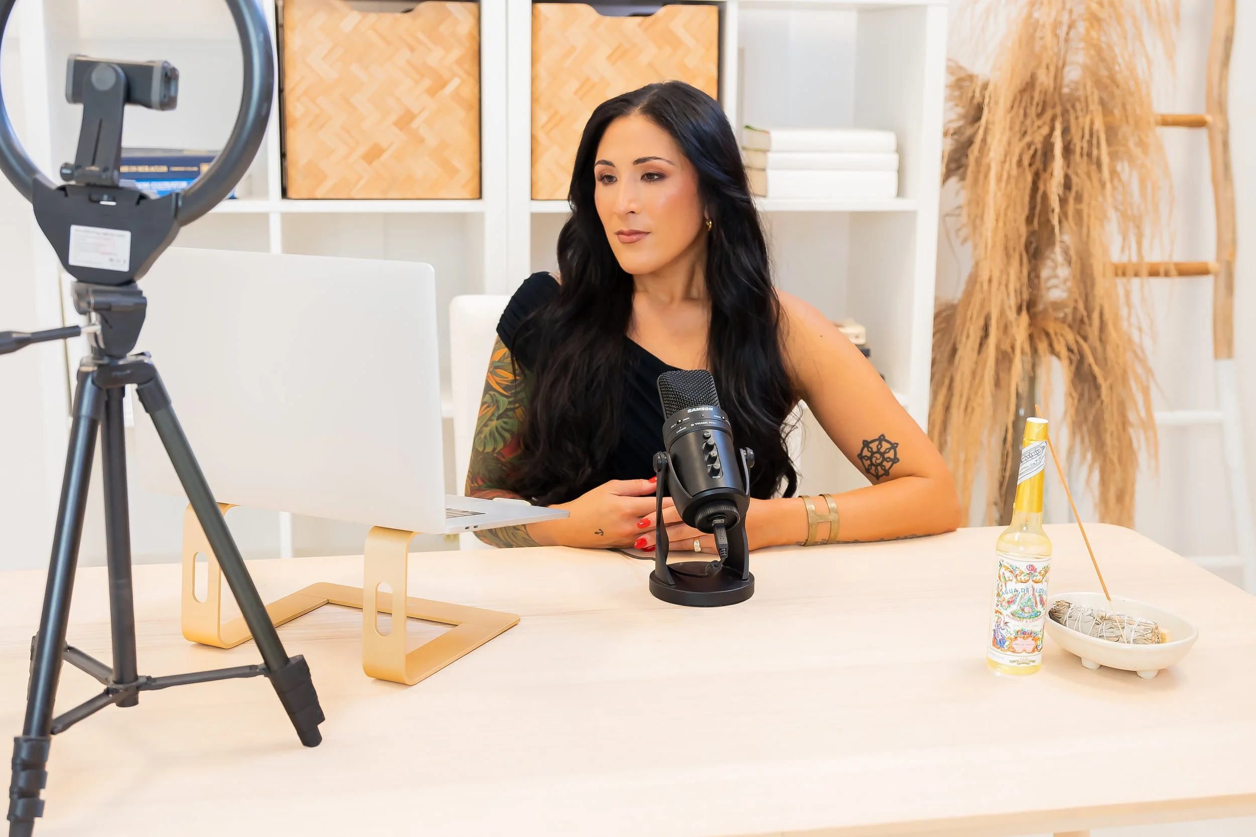 A woman with long black hair and colorful tattoos on her arms, sitting at a desk with a computer, microphone, and camera setup for recording or streaming, in a minimalist room with decorative plants and shelves.