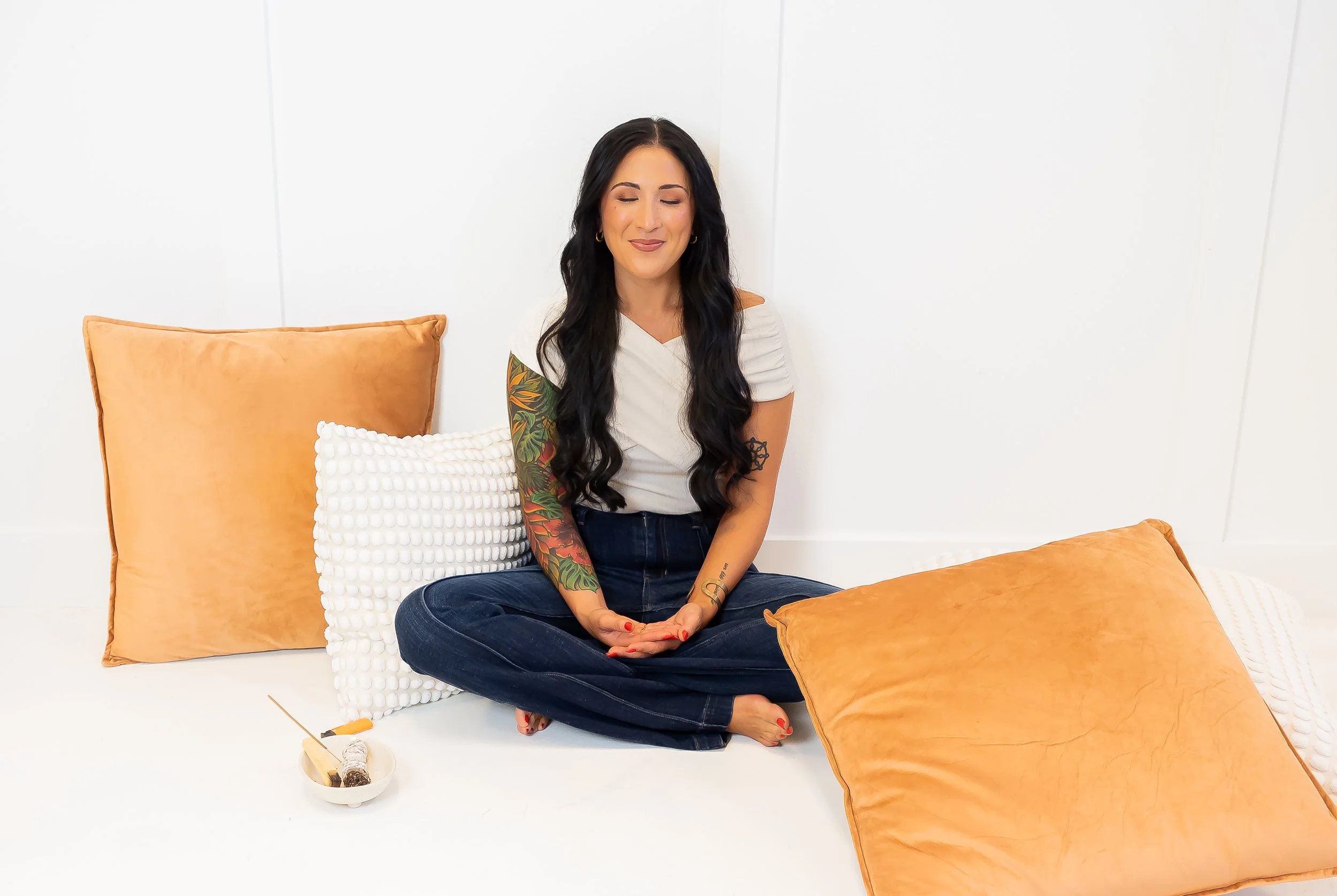 A woman with long dark hair and tattoos sitting cross-legged on the floor, surrounded by large throw pillows in shades of orange and white, with a bowl containing incense and a small stick nearby, against a plain white background.