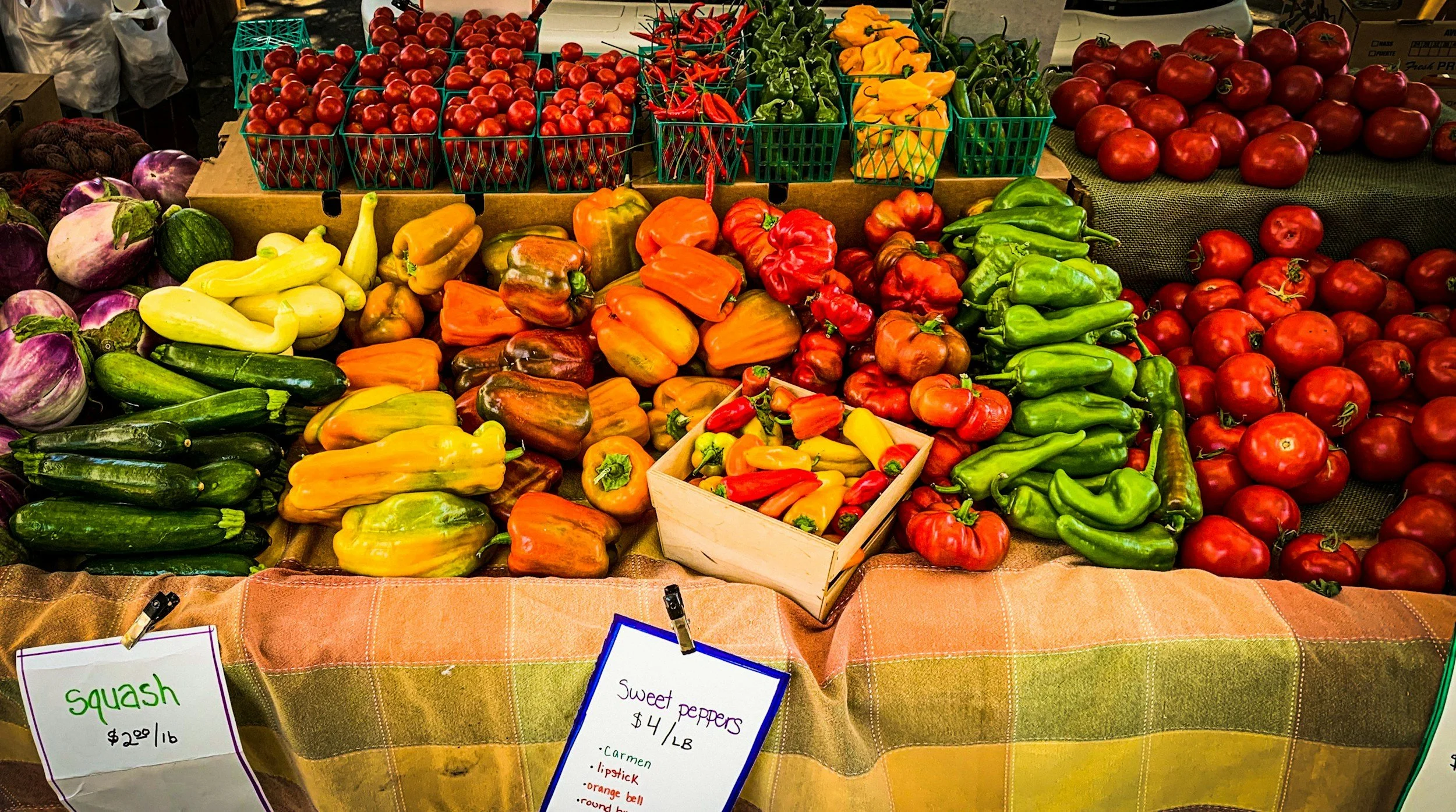 Display of fresh vegetables at a market stall, including tomatoes, bell peppers, squash, cucumbers, and chili peppers, with handwritten price signs.